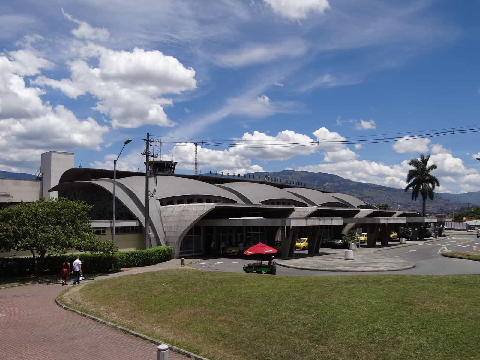 Frontansicht des Flughafens Olaya Herrera in Medellín: historisches Gebäude brutalistischer Architektur und Nationaldenkmal, mit Kontrollturm im Hintergrund, blauem Himmel und Palmen am Fußgängerzugang