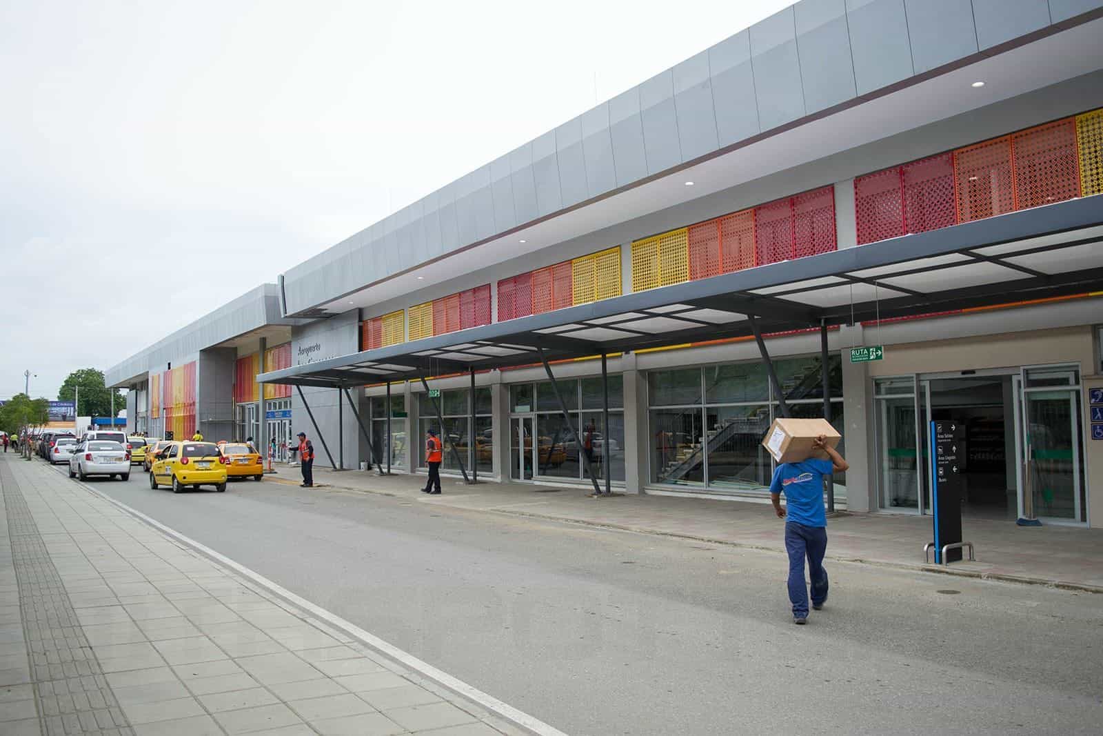 Hauptfassade des Internationalen Flughafens Los Garzones in Montería in der Abenddämmerung: einstöckige weiße Architektur mit großen Firmenbuchstaben „AEROPUERTO LOS GARZONES" über dem Fußgängereingang, geschwungenes Metallregendach über dem Ankunfts- und Taxibereich, Palmen zu beiden Seiten und klarer Himmel der kolumbianischen Karibik