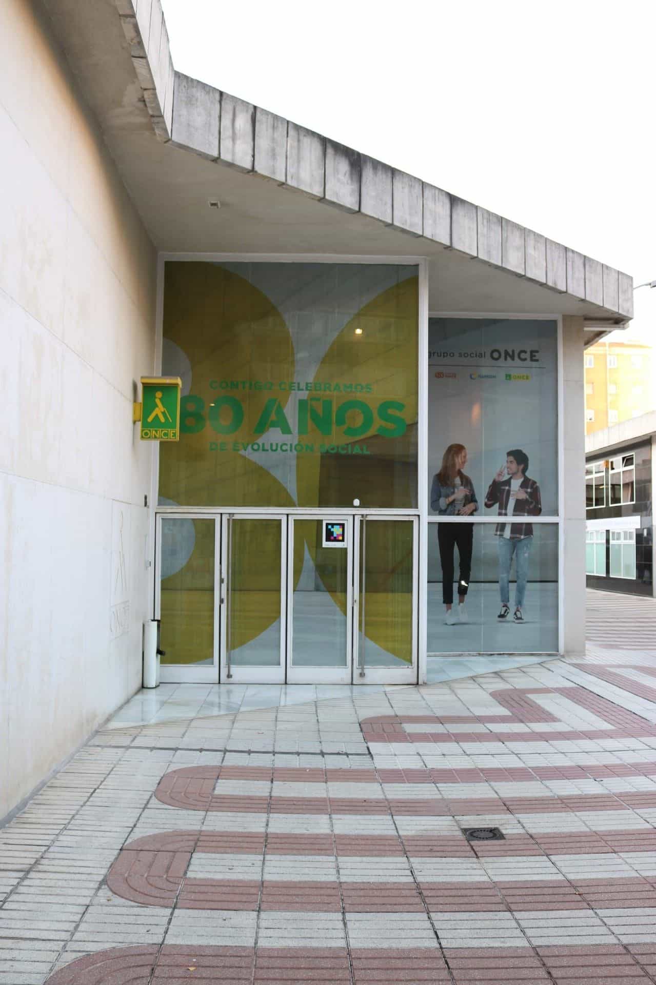 Facciata della Delegazione territoriale dell’ONCE a Valladolid: grande vetrata con il telone commemorativo «Contigo celebramos 80 años de evolución social» e il logo del Grupo Social ONCE; accanto alla porta d’ingresso, un codice NaviLens quadrato a colori e, di fianco, l’insegna giallo e verde del logo dell’ONCE con il bastone bianco