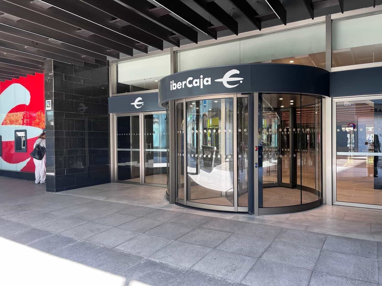 Fachada lateral de la sede central de Ibercaja en Zaragoza con la puerta giratoria de cristal bajo el rótulo «iberCaja» y, a la izquierda, un cajero rojo y un código NaviLens pegado en el pilar de granito negro junto al cajero