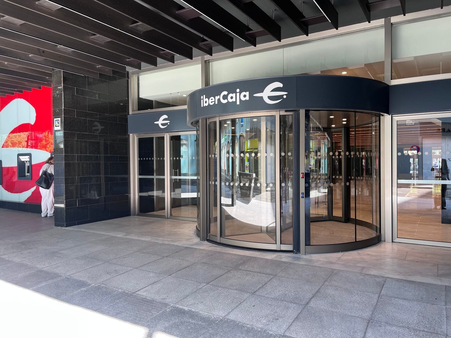 Fachada lateral de la sede central de Ibercaja en Zaragoza con la puerta giratoria de cristal bajo el rótulo «iberCaja» y, a la izquierda, un cajero rojo y un código NaviLens pegado en el pilar de granito negro junto al cajero