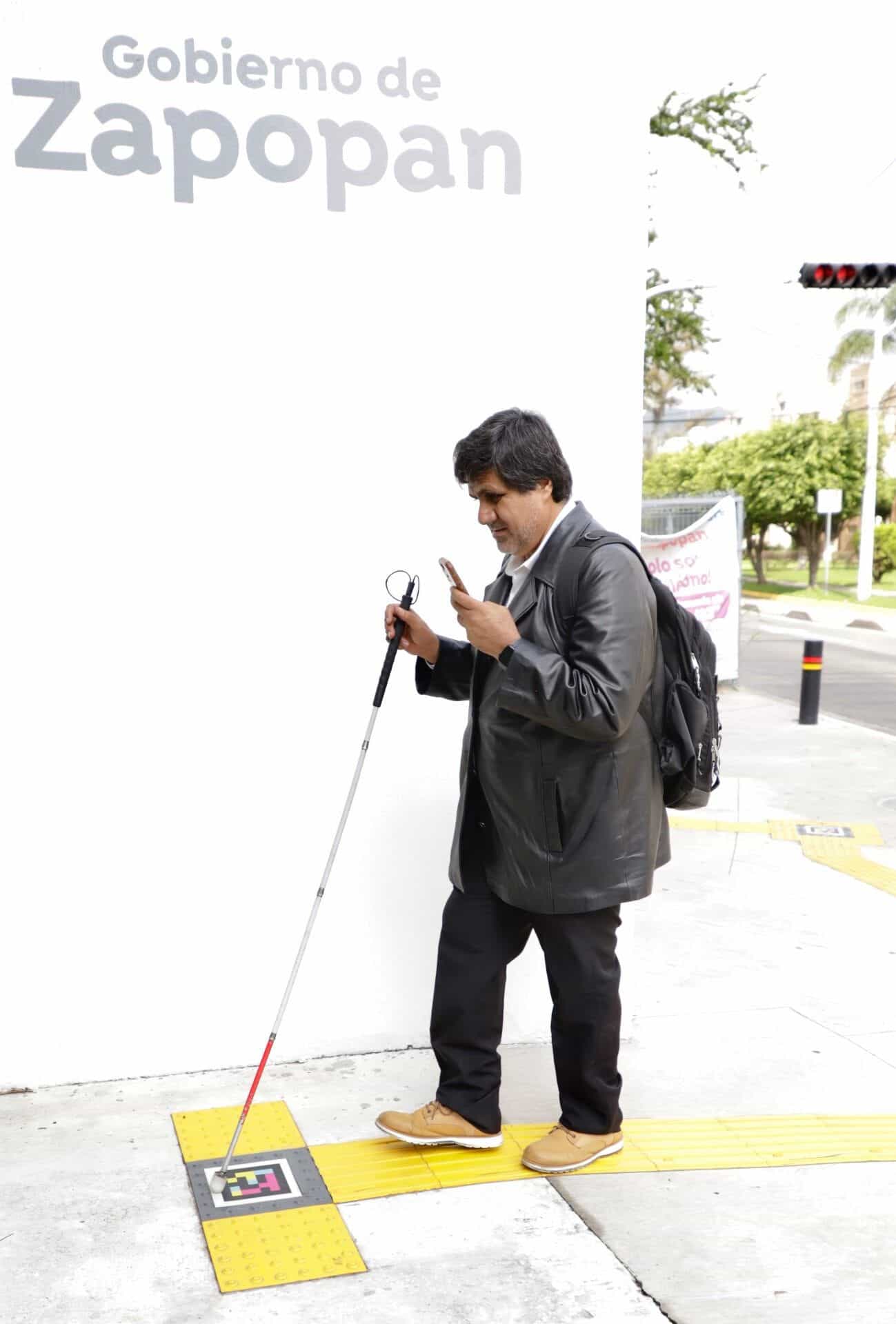 User with a white cane and a phone in front of the Zapopan Government building, next to a NaviLens code embedded in the yellow tactile mosaic of the sidewalk