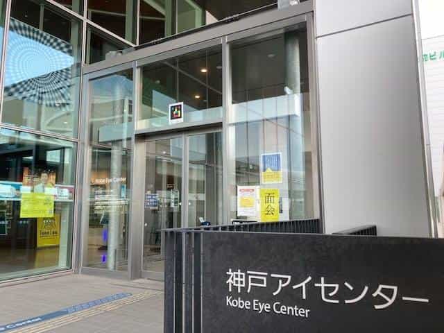 Glass façade of the Kobe Eye Center with a NaviLens tag visible above the entrance and a black totem with the “神戸アイセンター / Kobe Eye Center” sign in the foreground