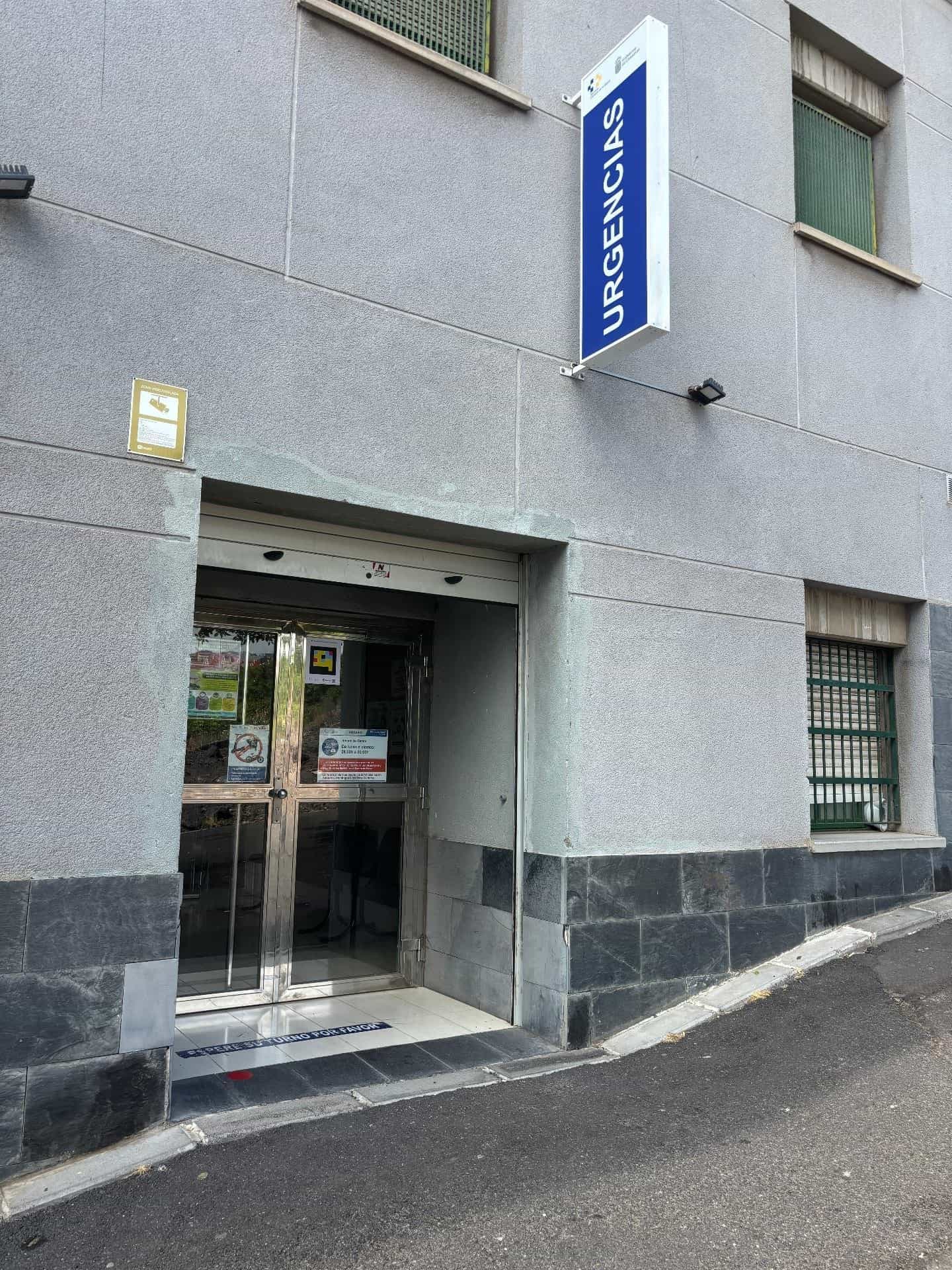 テネリフェのカナリア保健サービス保健センターのファサード:黒い石の腰壁付きの淡灰色の壁、白文字で「URGENCIAS」(救急)と書かれた青い照明付き庇、金属枠のガラス扉、左側の扉にはセンターの他の通知と並んでNaviLensコードの掲示物。床には「ESPERE SU TURNO POR FAVOR」と書かれている。