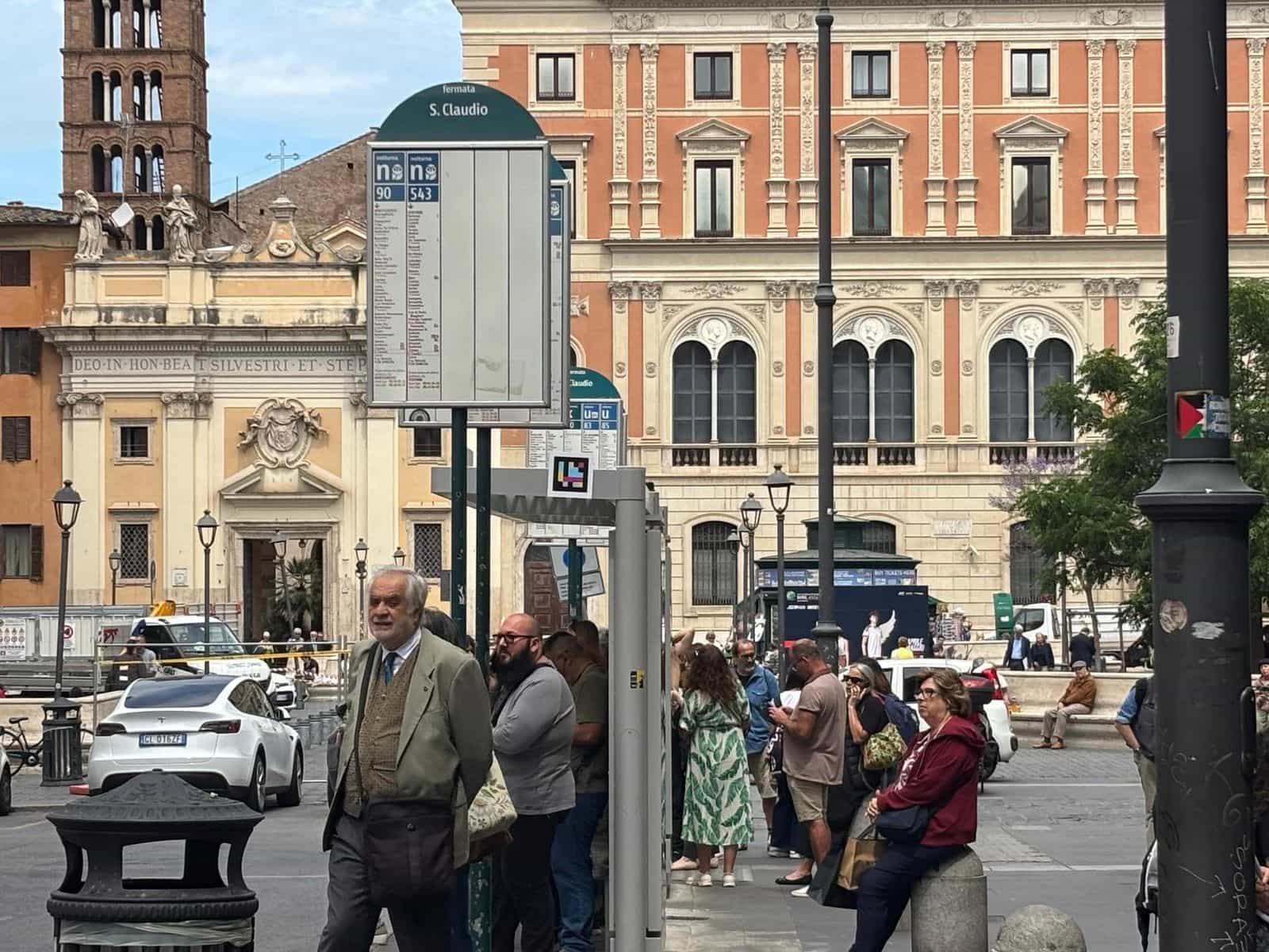Piazza S. Claudio a Roma con il cartello Bus info di ATAC e NaviLens visibile sul vetro della pensilina, davanti a una chiesa barocca