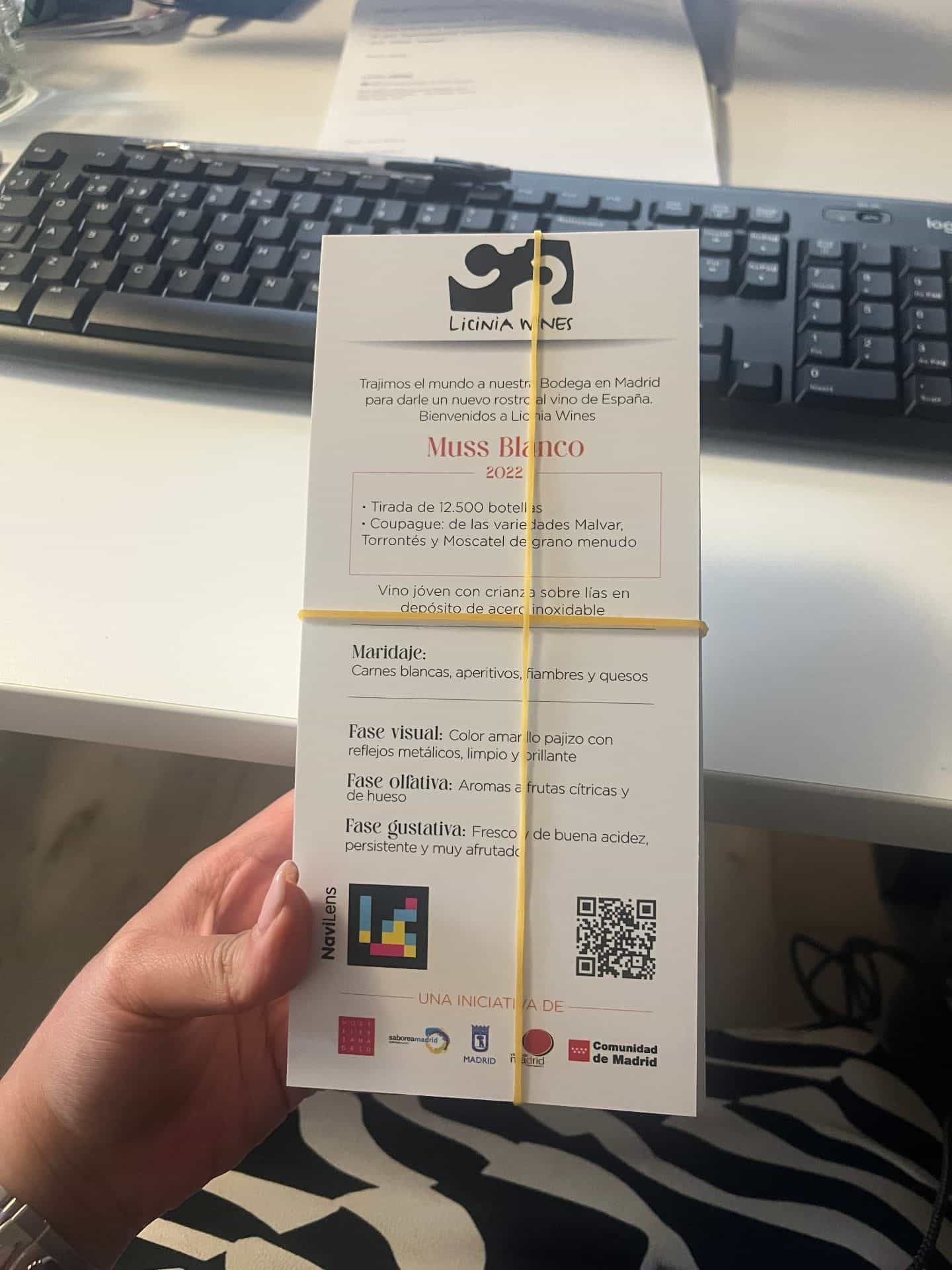 Scheda di degustazione di Decanta Madrid tenuta con un elastico giallo: in alto il logo «Licinia Wines» con un tappo disegnato; scheda del vino «Muss Blanco 2022» con tiratura, cuvée di Malvar, Torrontés e Moscato a grana piccola, abbinamento e fasi visiva, olfattiva e gustativa; in basso a sinistra un codice NaviLens quadrato a colori con il marchio «NaviLens», a destra un QR nero e, in fondo, i logo «UNA INICIATIVA DE» Hostelería Madrid, Saborea Madrid, Comune di Madrid e Comunità di Madrid