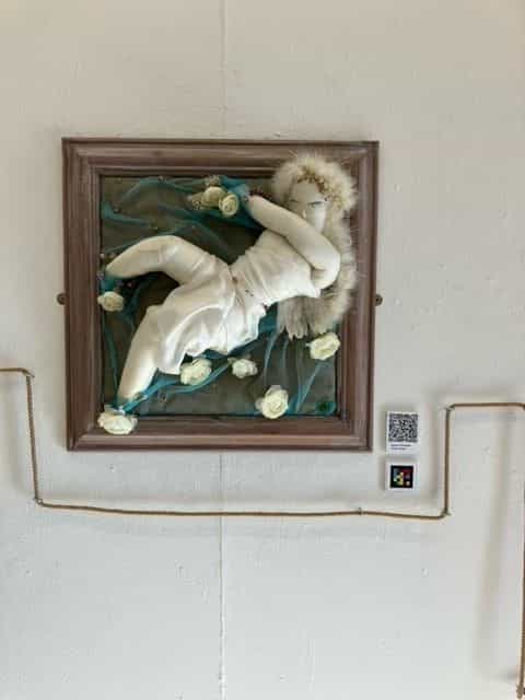 Opera incorniciata in legno scuro su parete bianca: dentro la cornice, su tela increspata verde-azzurra, una figura femminile scultorea di stoffa bianca con vestito e velo di piume, circondata da piccole rose bianche; a destra della cornice, una piccola didascalia bianca con codice NaviLens e un codice QR