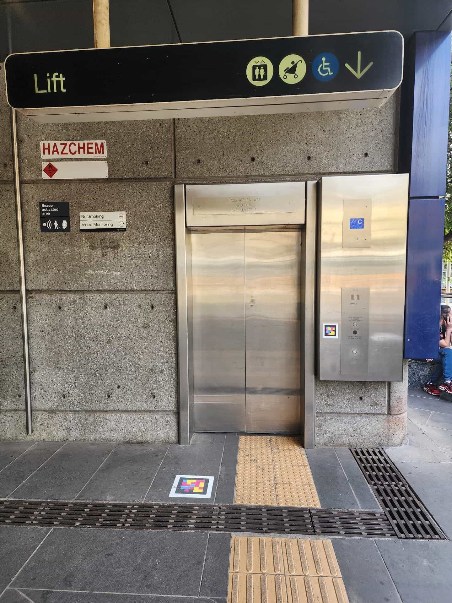 Accesso al «Lift Number 2» della Flagstaff Railway Station di Melbourne: cartello nero sospeso con la parola «Lift» in verde e i pittogrammi di ascensore, passeggino e sedia a rotelle; ascensore metallico in acciaio inox con pulsantiera laterale su cui si nota un codice NaviLens con cornice bianca accanto al pulsante, su pavimento di granito grigio con fascia tattile gialla