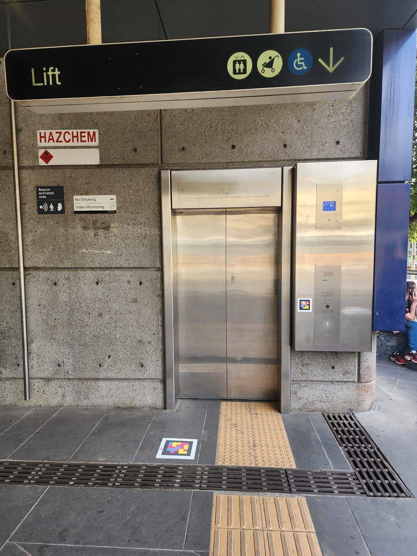 Access to “Lift Number 2” at Melbourne's Flagstaff Railway Station: black overhead sign reading “Lift” in green with the lift, pram and wheelchair pictograms; stainless steel lift with a side panel showing a white-framed NaviLens code next to the button, on grey granite flooring with a yellow tactile strip