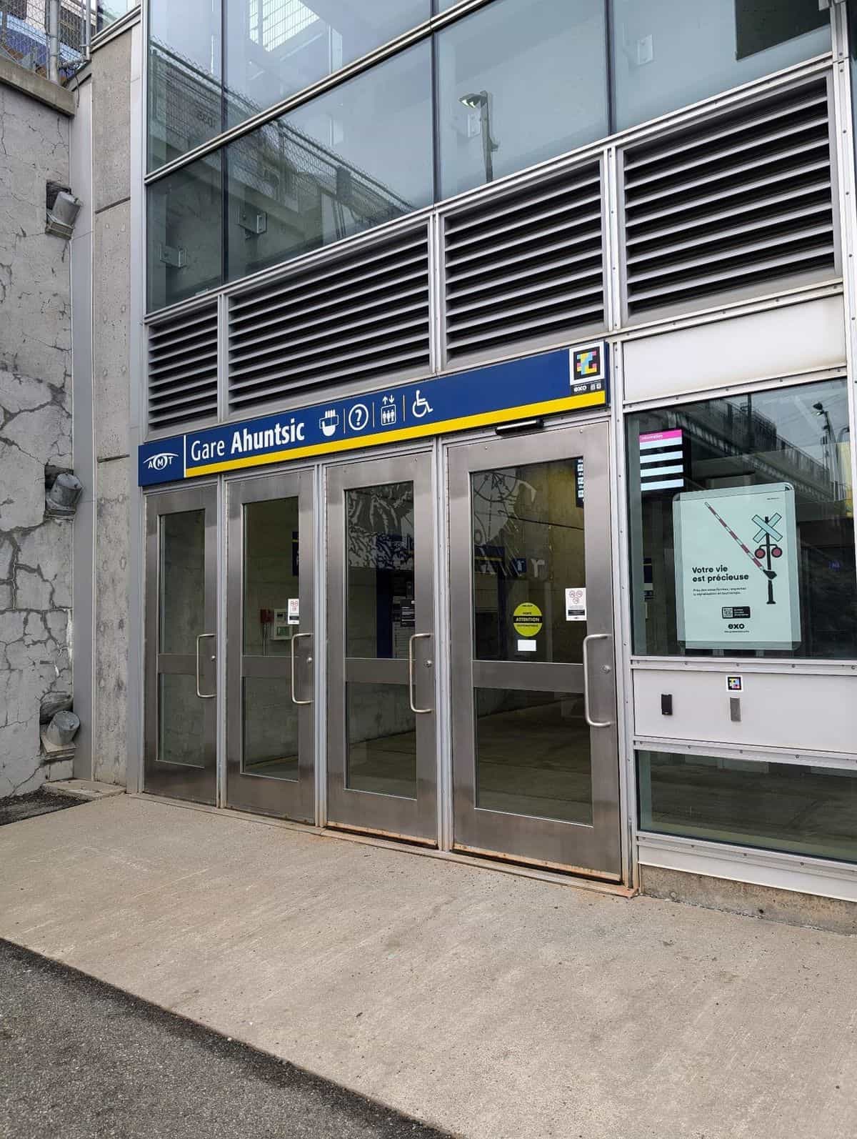 Main entrance of exo's Gare Ahuntsic in Montréal: four metal-framed glass doors below a large blue-and-yellow «Gare Ahuntsic» sign with the AMT logo and pictograms for café, info, elevator and wheelchair; in the upper-right corner, the NaviLens code in cyan, magenta and yellow on a black background, with the exo logo just below