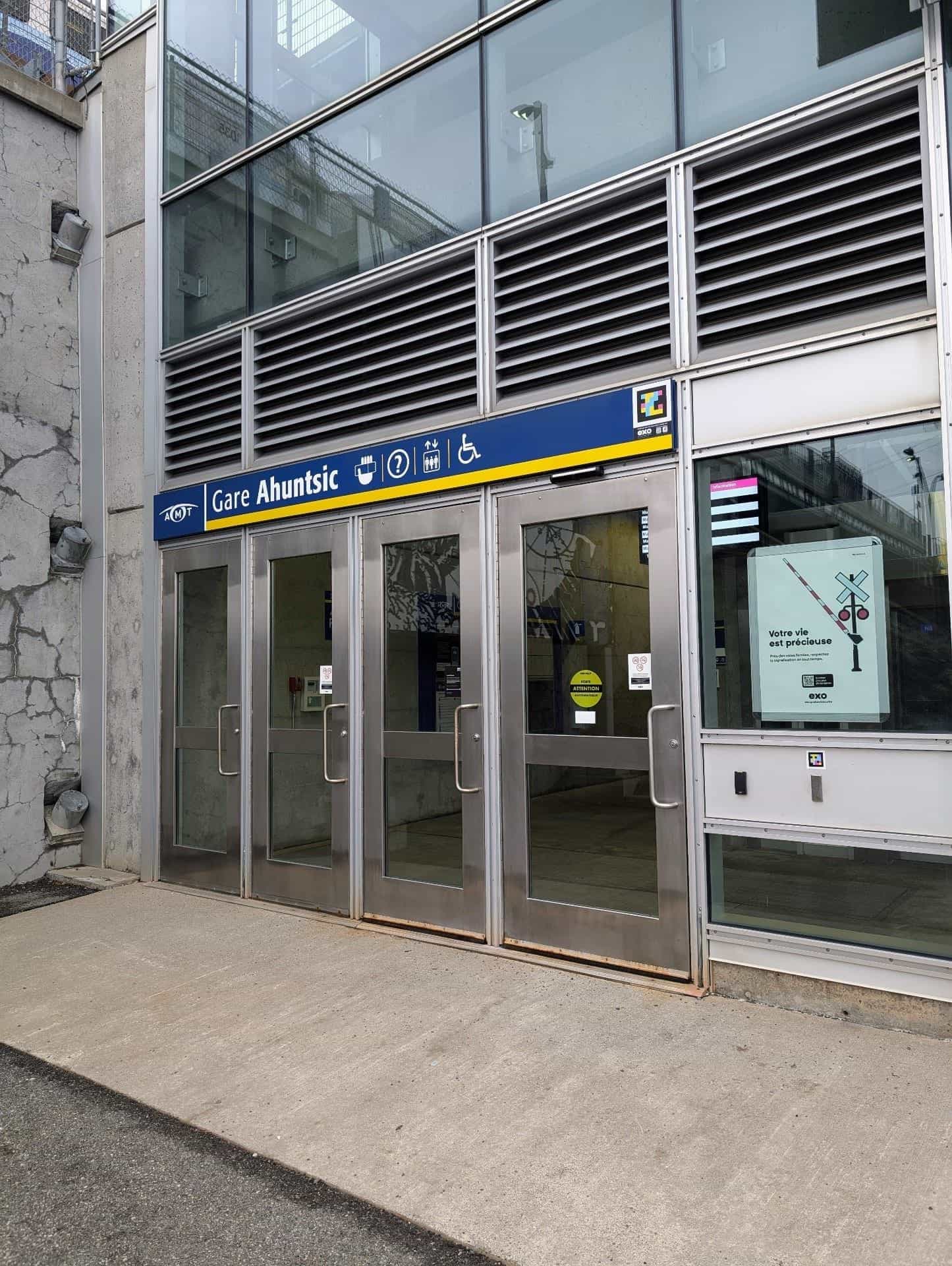 Main entrance of exo's Gare Ahuntsic in Montréal: four metal-framed glass doors below a large blue-and-yellow “Gare Ahuntsic” sign with the AMT logo and pictograms for café, info, elevator and wheelchair; in the upper-right corner, the NaviLens code in cyan, magenta and yellow on a black background, with the exo logo just below