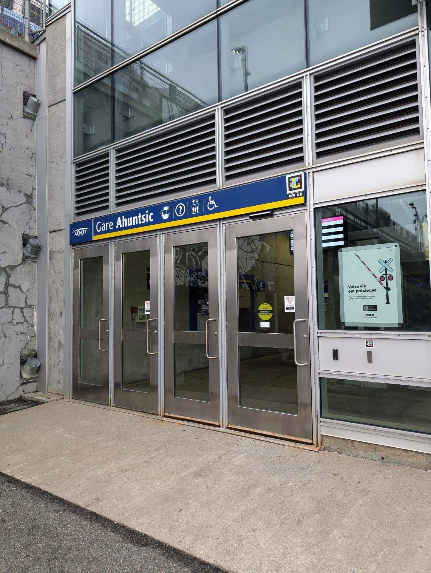 Main entrance of exo's Gare Ahuntsic in Montréal: four metal-framed glass doors below a large blue-and-yellow “Gare Ahuntsic” sign with the AMT logo and pictograms for café, info, lift and wheelchair; in the upper-right corner, the NaviLens code in cyan, magenta and yellow on a black background, with the exo logo just below