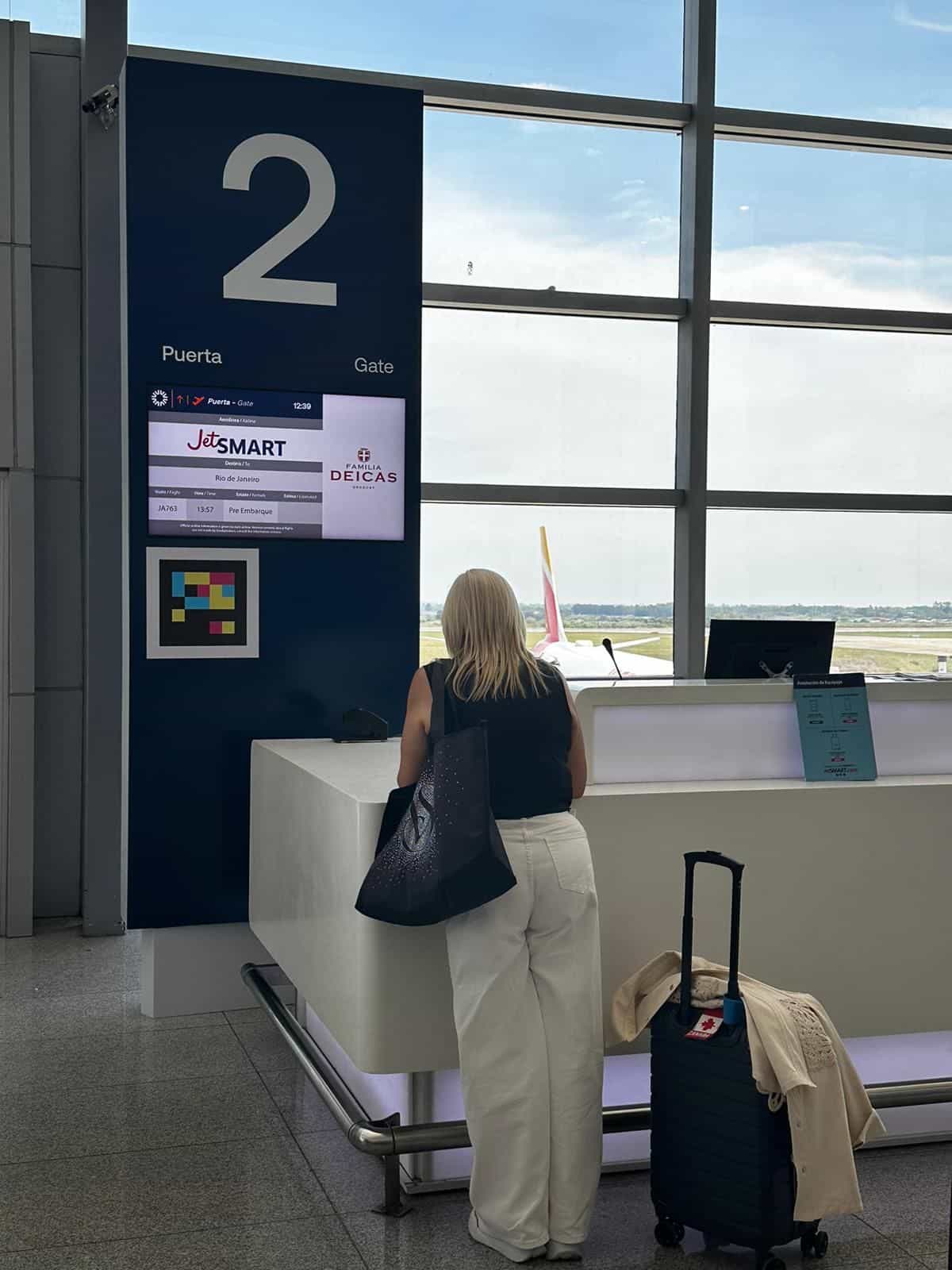Schalter am Puerta 2 / Gate 2 des Flughafens Carrasco: große marineblaue Säule mit weißem «2», Aufschrift «Puerta · Gate», FIDS-Bildschirm «JetSMART · Rio de Janeiro · JA763 · 13:57 · Pre-Boarding», darunter ein quadratischer NaviLens-Code in Cyan, Magenta, Gelb und Rot mit weißem Rahmen