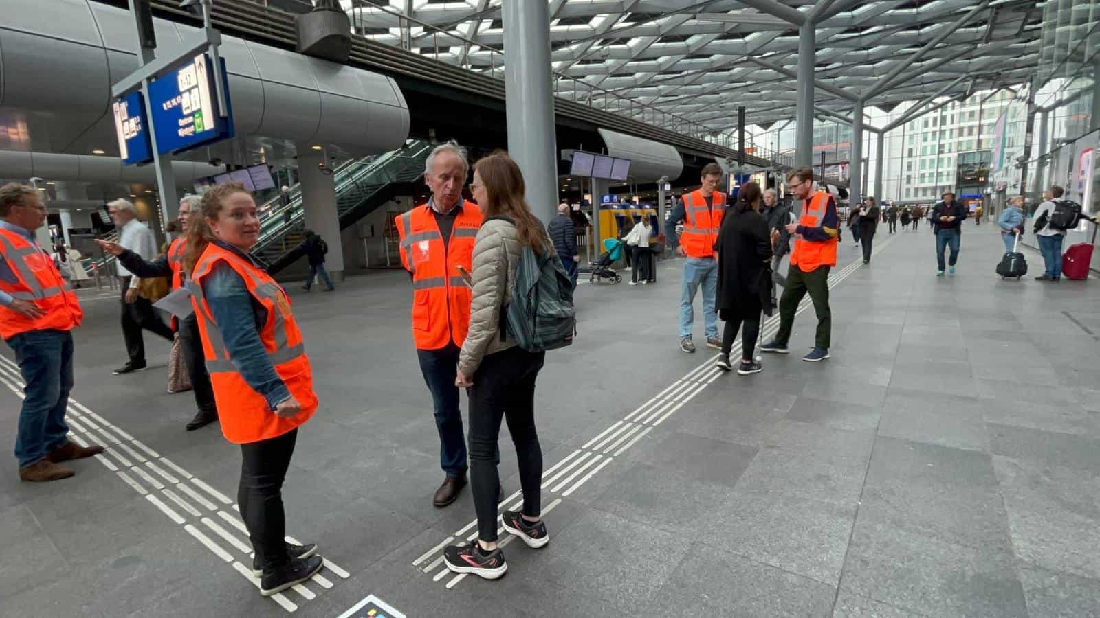 ProRail team in orange vests and volunteers standing on the tactile guide line in the main hall of Den Haag Centraal during the indoor navigation trial of June 2022, with a NaviLens code visible on the pavement
