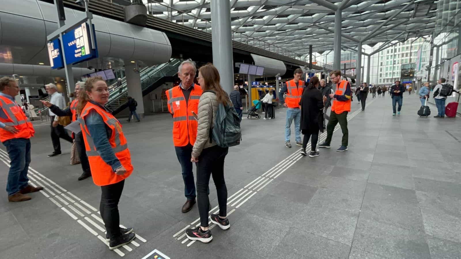 Squadra di ProRail con gilet arancioni e persone volontarie in piedi sulla linea guida tattile dell’atrio principale di Den Haag Centraal durante il test di navigazione indoor del giugno 2022, con un codice NaviLens visibile sul pavimento