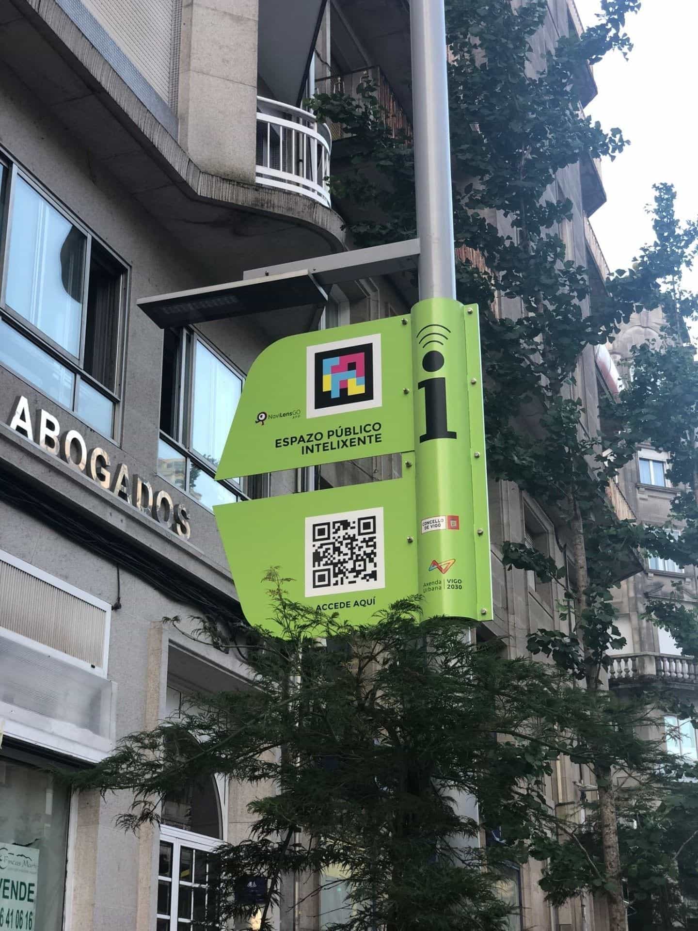 Primo piano della targa verde dell’Espazo Público Intelixente appesa a un palo della luce nella Gran Vía di Vigo, accanto a un’insegna «ABOGADOS»: il pannello mostra il logo NaviLens GO, un codice NaviLens quadrato a colori, la scritta «Espazo Público Intelixente», un QR «Accede aquí» e gli emblemi del Comune di Vigo e dell’Axenda Urbana Vigo 2030