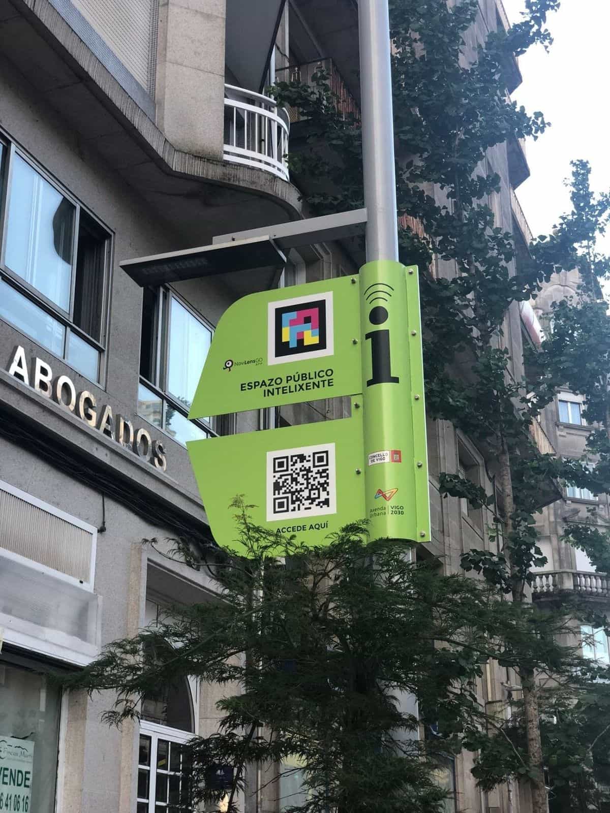 Plano detalle de la placa verde del Espazo Público Intelixente colgada de una farola en la Gran Vía de Vigo, junto a un letrero 'ABOGADOS': el panel muestra el logotipo NaviLens GO, un código NaviLens cuadrado a colores, 'Espazo Público Intelixente', un QR 'Accede aquí' y los emblemas del Concello de Vigo y la Axenda Urbana Vigo 2030