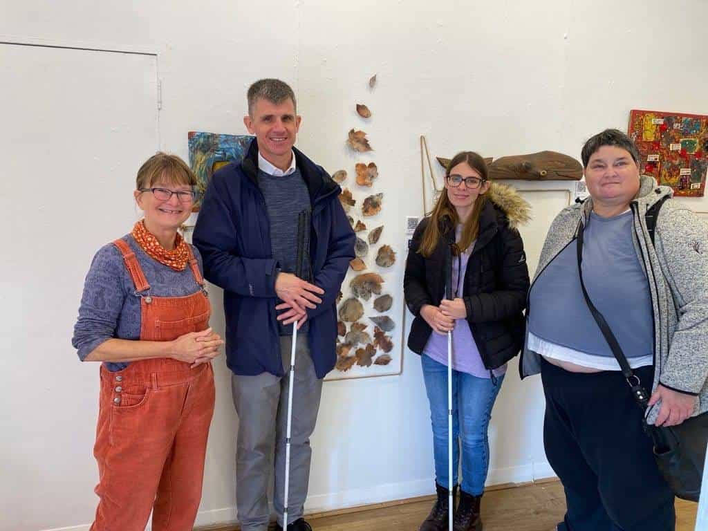 Group of four people smiling in front of the white gallery wall, two of them holding white canes, beside artists from the collective with several works in the background