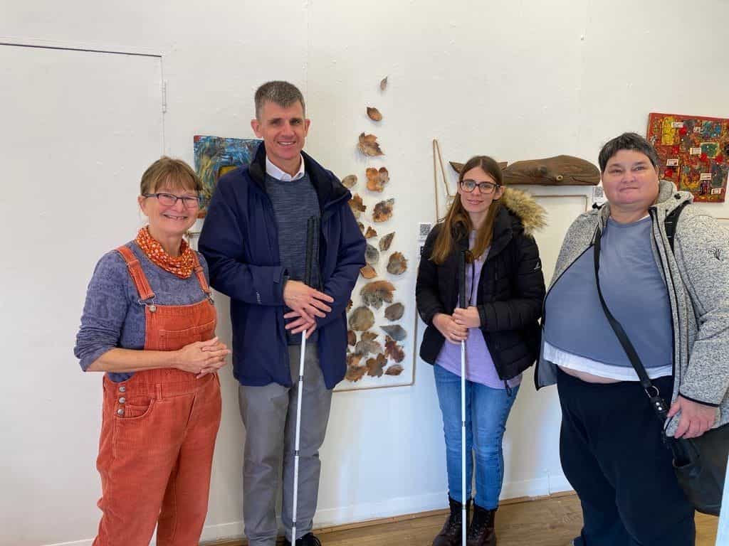 Gruppo di quattro persone che posano sorridenti nella galleria davanti alla parete bianca, due di loro con bastone bianco, accanto ad artisti del collettivo e a diverse opere in fondo