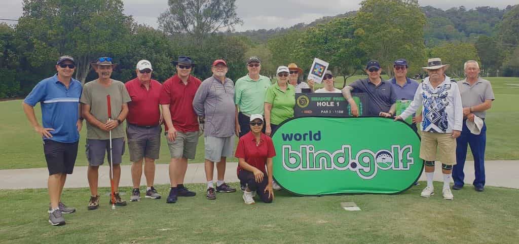 Gruppo di golfisti e accompagnatori in posa davanti al cartello della Buca 1 di Meadow Park, con lo stendardo verde di World Blind Golf e la targa Tee See con codice NaviLens