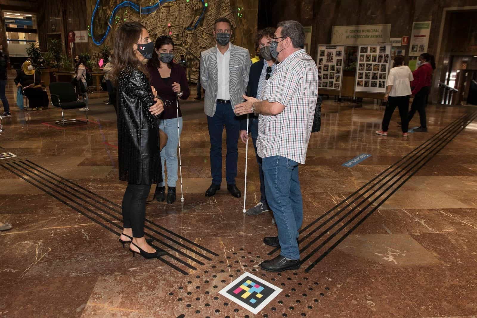 Gruppo di persone con la mascherina nella hall del Comune di Saragozza, accanto a un’utente della ONCE con bastone bianco; sul pavimento in marmo, fasce tattili nere che convergono in una croce e un codice NaviLens quadrato a colori applicato al centro; sullo sfondo, il grande plastico-murale in rilievo della città di Saragozza con l’Ebro illuminato di blu