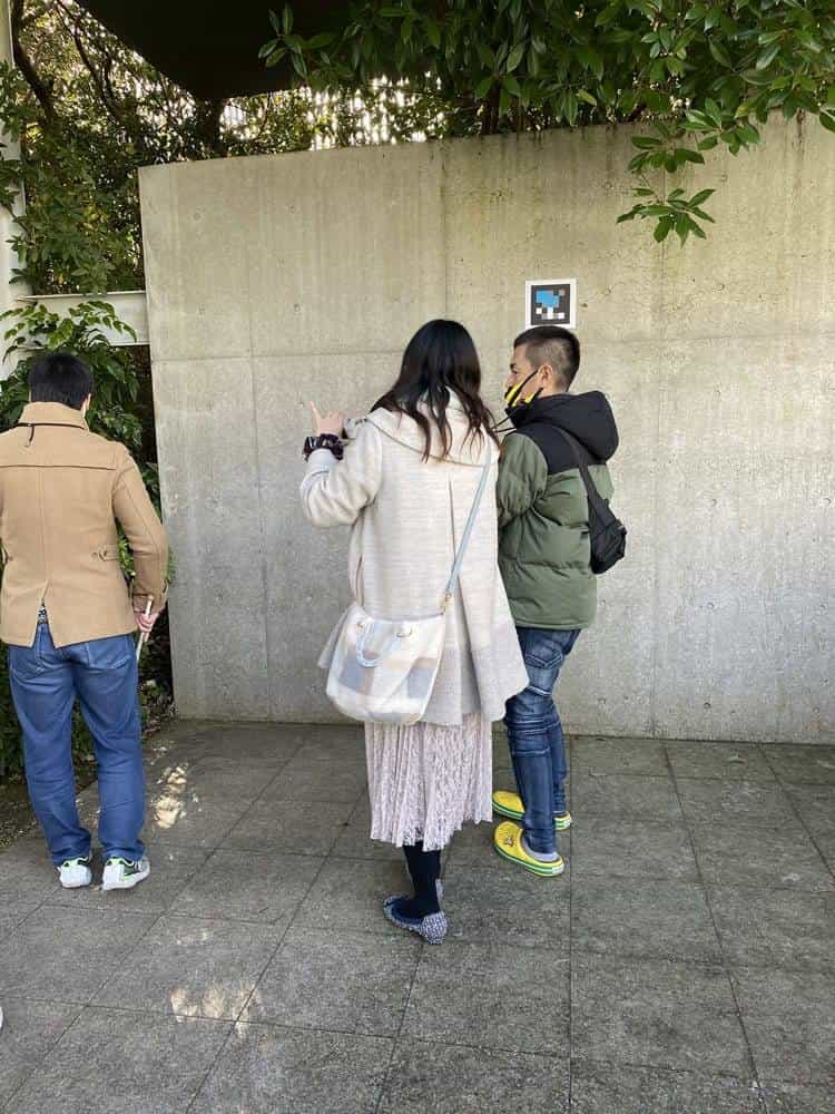 Group of three visitors – one man and two young people – in front of a concrete wall with a NaviLens code, one of them pointing at the code