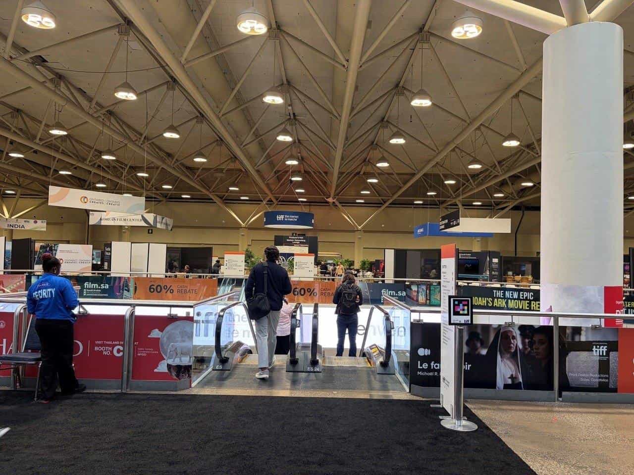 The floor of the Metro Toronto Convention Centre during TIFF: The Market. Under the “tiff · the market · le marché” banner hanging from the ceiling, two delegates ride the escalators down past the Ontario Creates, film.sa and Korean Film Center booths; on the right, a NaviLens code mounted on a stanchion beside the film posters.