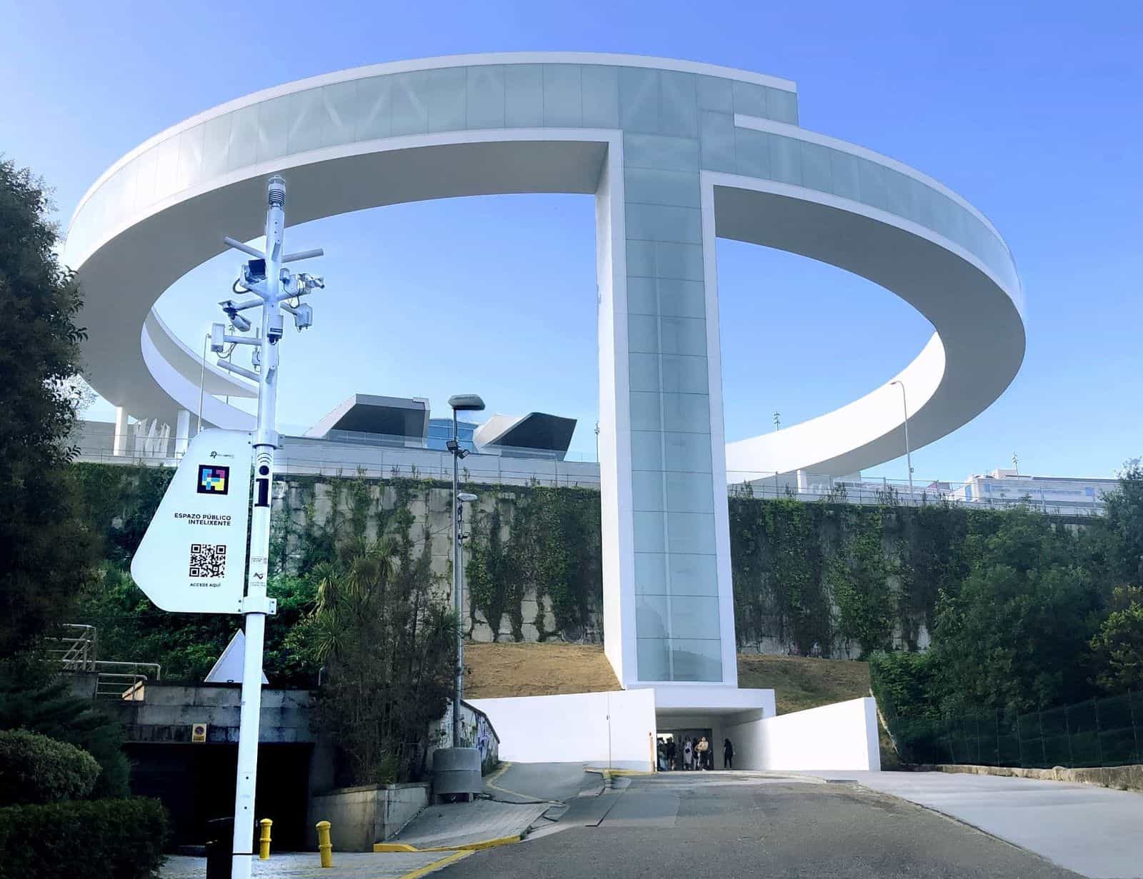 Accesso inferiore all’ascensore HALO di Vigo in Serafín Avendaño: grande struttura bianca circolare sopra il muro vegetale, con la passerella a forma di anello e l’ascensore a vetri; in primo piano, un palo di smart city con telecamere e sensori IOTSENS e una targa bianca con il codice NaviLens GO «Espazo Público Intelixente · Accede aquí» del Comune di Vigo