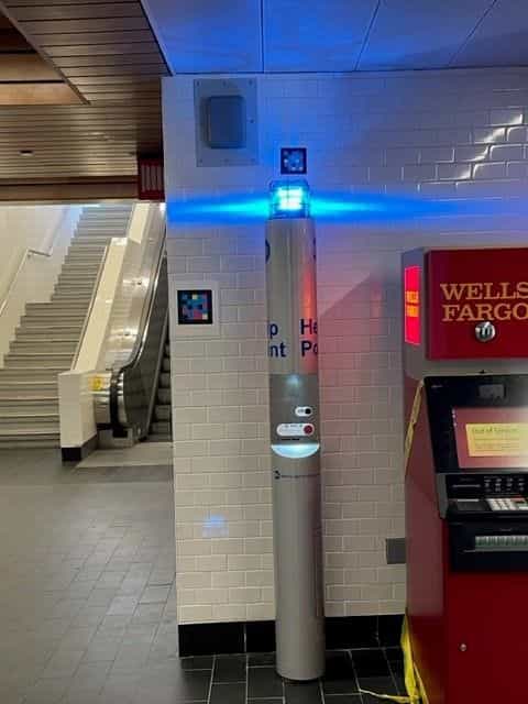 Blue illuminated Help Point at White Plains station, next to a white-framed NaviLens code on the tiled wall; escalators in the background