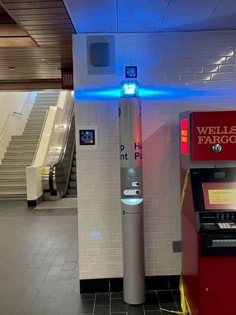 Blue illuminated Help Point at White Plains station, next to a white-framed NaviLens code on the tiled wall; escalators in the background