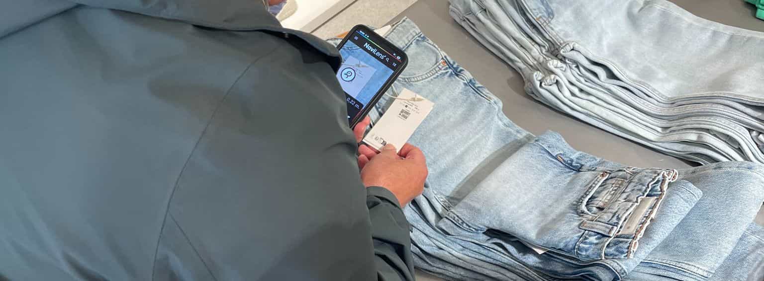 Person scannt das Hangtag einer Jeans mit der NaviLens-App; der NanoLens-Code ist neben dem QR-Code von zara.com gedruckt.