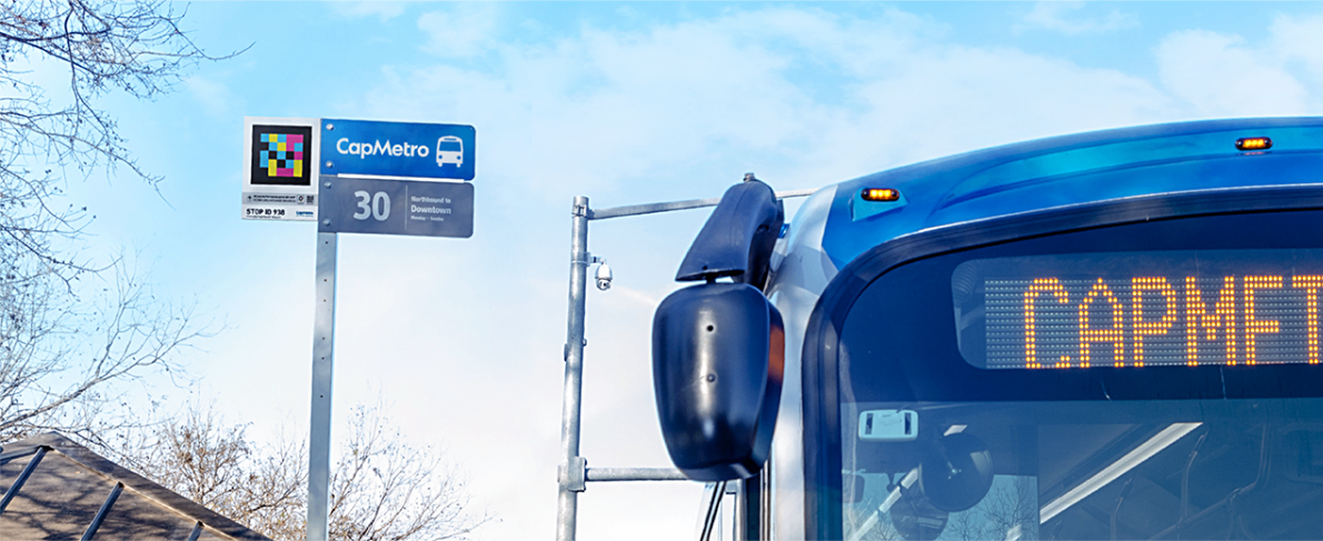 Parada de CapMetro (Austin) con un código NaviLens junto al logo en el poste; a la derecha, el frontal de un autobús azul con cartel LED.