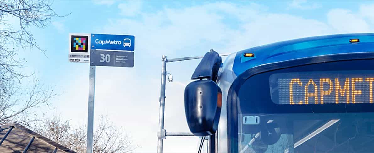 Fermata di CapMetro (Austin) con un codice NaviLens accanto al logo sulla palina; a destra, il frontale di un autobus blu con display LED.