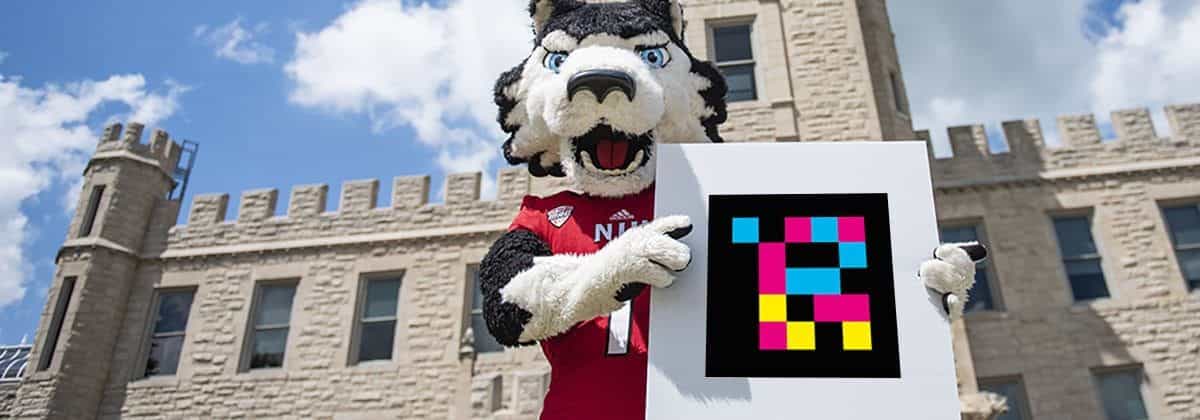 Victor E. Huskie, Maskottchen der Northern Illinois University, hält einen NaviLens-Code vor dem Castle-Gebäude der NIU