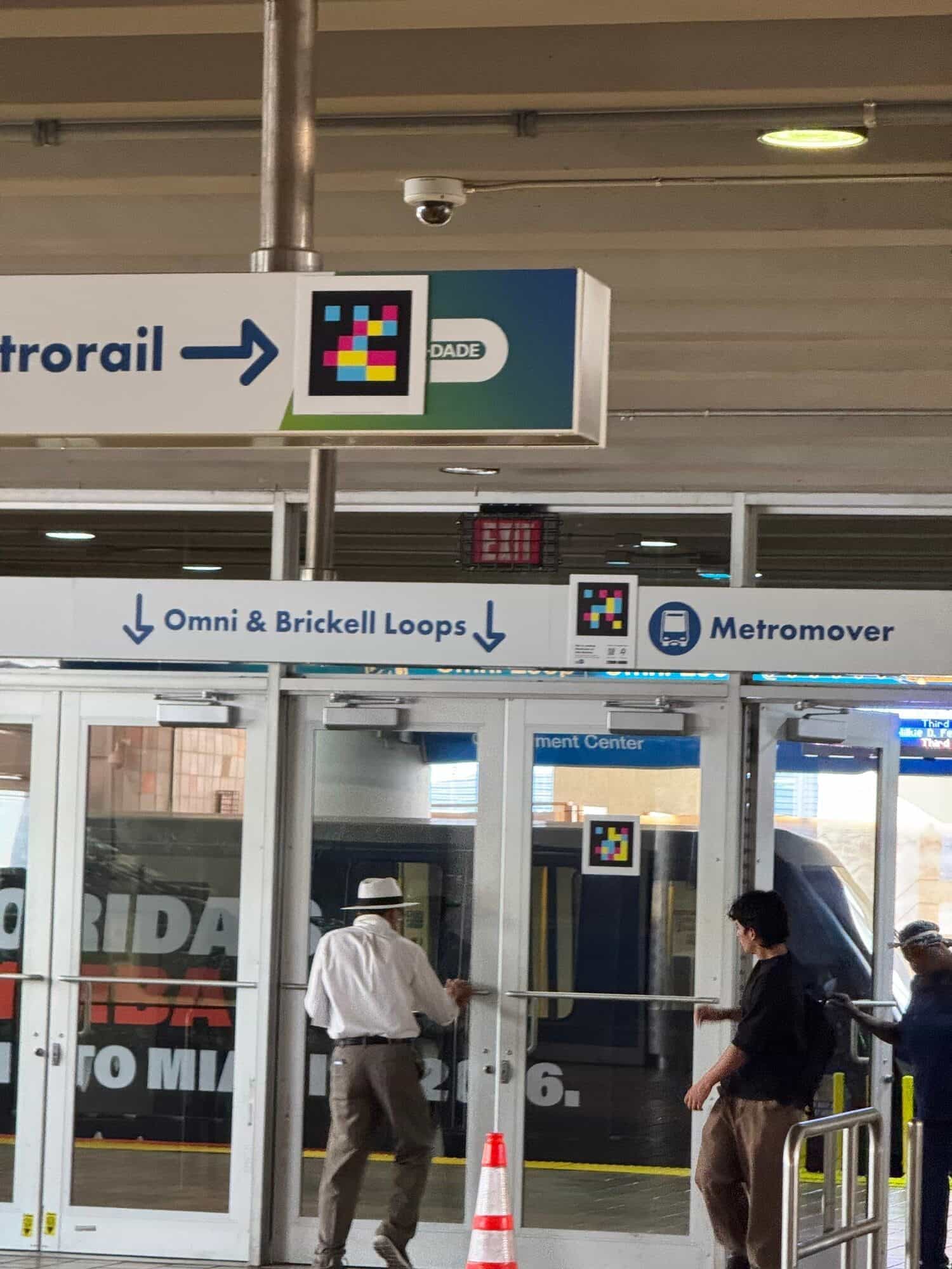 Segnaletica sospesa di Miami-Dade Metrorail con freccia azzurra «Metrorail →» e un codice NaviLens magenta, ciano e giallo integrato nel cartello sopra le porte verso gli Omni & Brickell Loops del Metromover