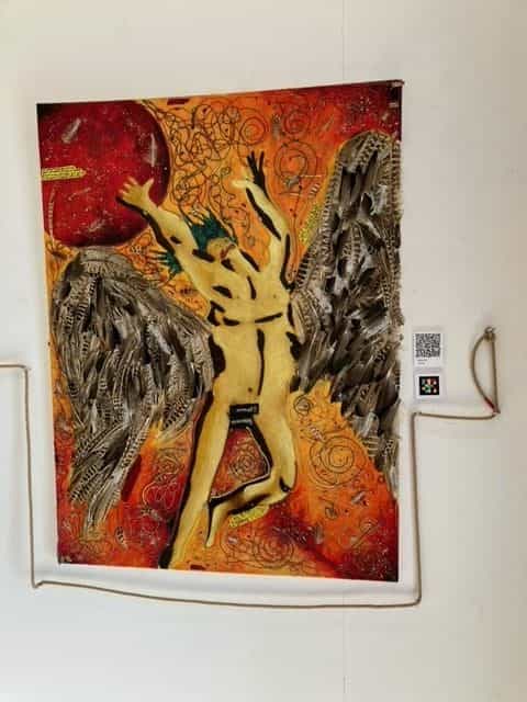 Opera tessile di grande formato appesa alla parete bianca: figura maschile dorata con le braccia aperte a croce e due enormi ali fatte di piume grigie e bianche vere su un fondo dipinto in arancione, giallo e rosso con la sagoma di un sole rosso nell’angolo superiore sinistro; a destra, sulla parete, una piccola didascalia bianca con codice NaviLens a quadretti gialli, rosa, azzurri e neri e un codice QR; in basso a destra, la corda guida di canapa tesa orizzontalmente