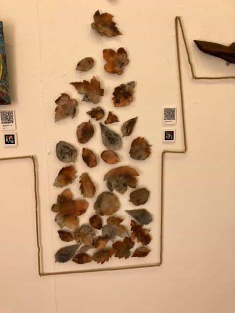 Cream gallery wall with a vertical composition of dried, rusted copper-and-grey leaves arranged into an elongated figure; around it, the hemp rope draws a stepped frame and two small white labels with NaviLens codes