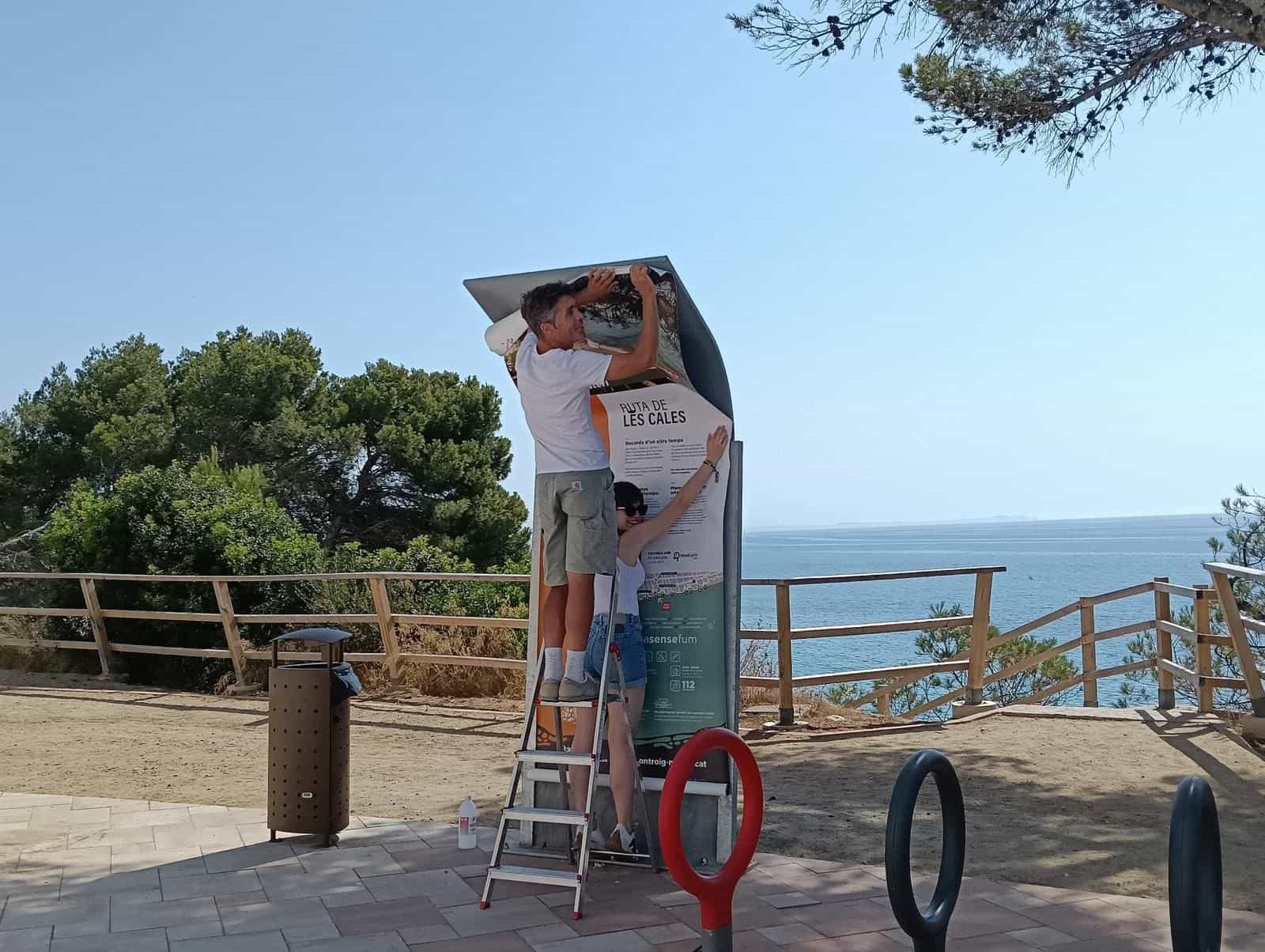 Zwei Personen bringen von einer Trittleiter die Grafikfolie eines Ruta-de-les-Cales-Totems an, auf einem Aussichtspunkt über dem Meer