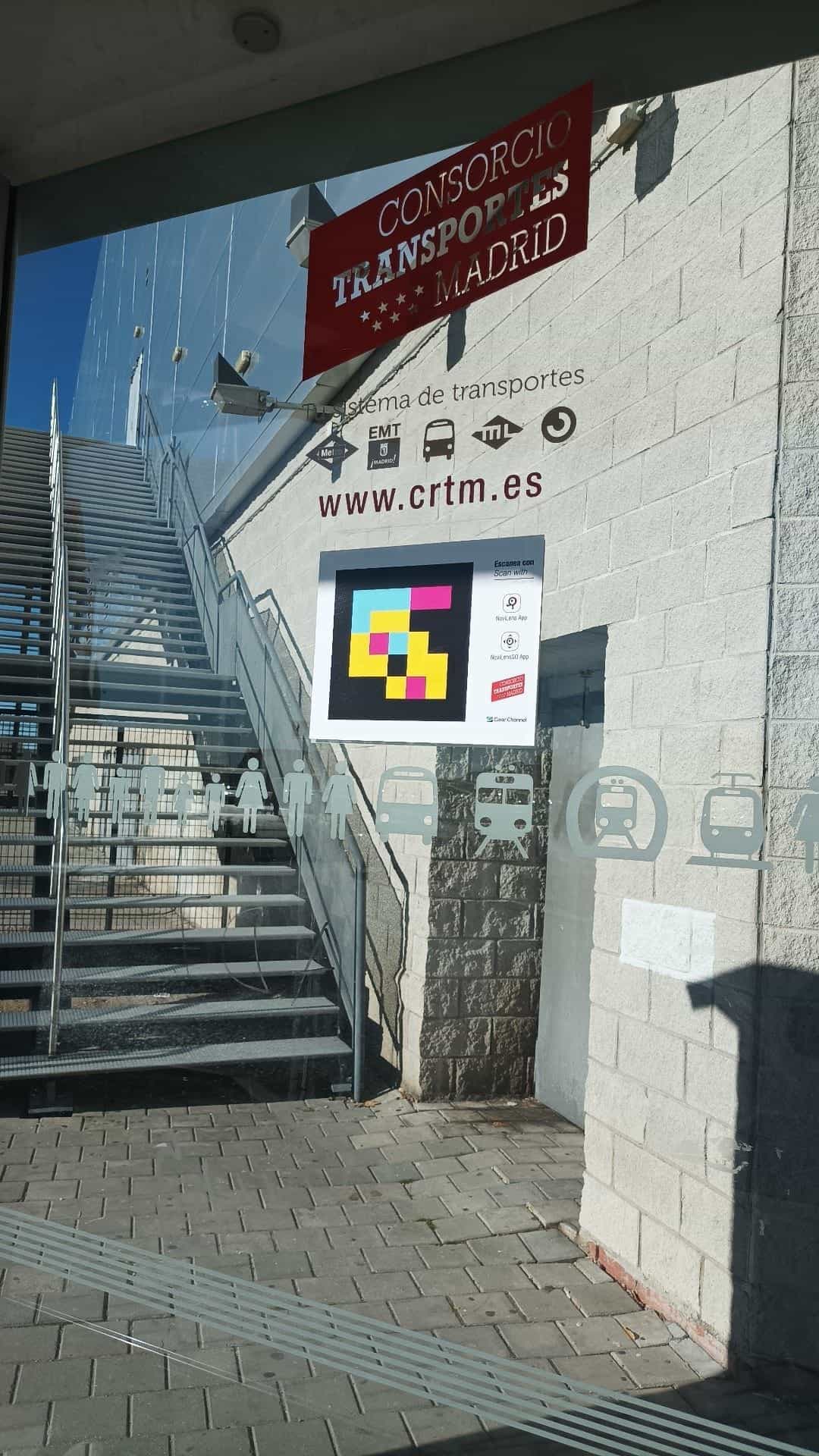 Cartel «Escanea con / Scan with» con un código NaviLens cuadrado a colores, iconos de las apps NaviLens y NaviLens GO, logo del CRTM y logo de Clear Channel pegado sobre el cristal de una marquesina