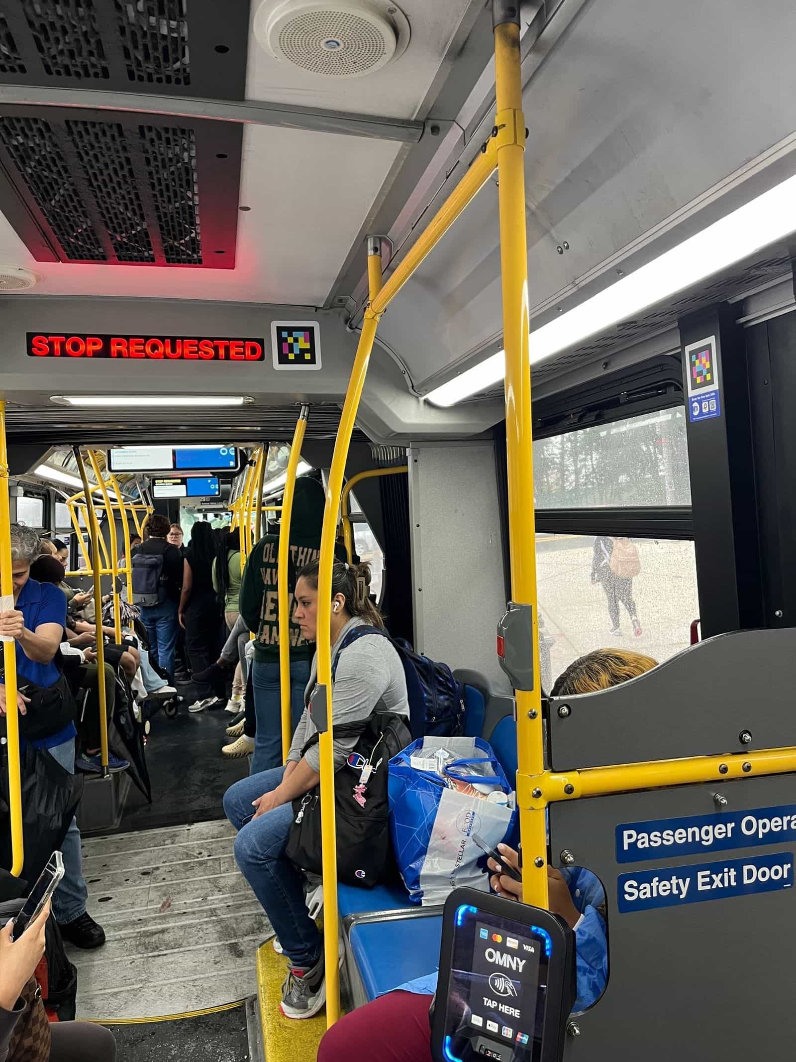 Interno del bus Bx12 con la scritta LED rossa «STOP REQUESTED» accesa e un codice NaviLens fissato subito a destra sul soffitto; un altro codice visibile sopra la porta posteriore accanto al lettore OMNY