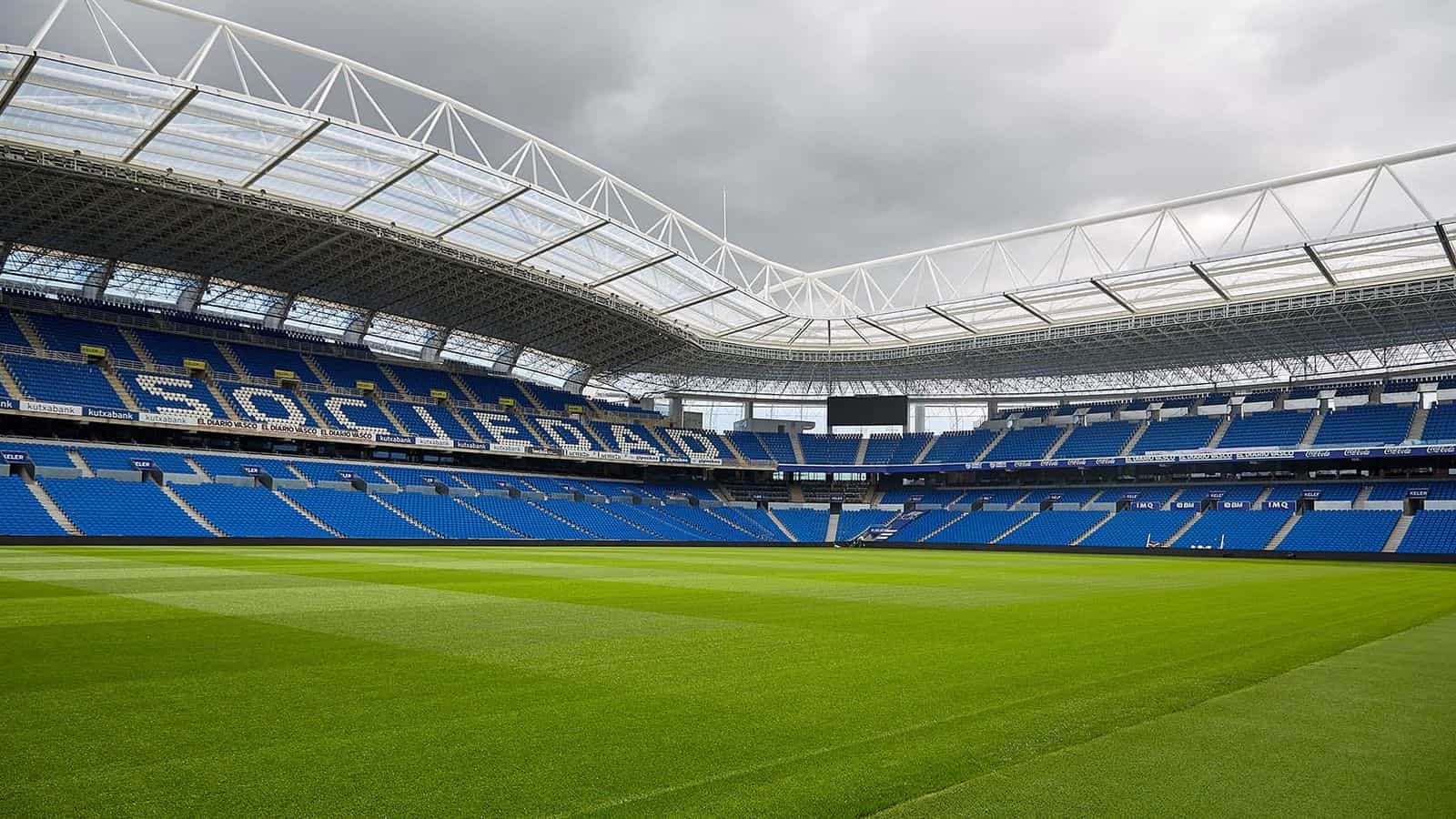 Innenraum des Reale Arena vom Spielfeld aus — blaue Tribünen, in deren Sitzen das Wort „SOCIEDAD“ ausgeschrieben ist, unter dem lichtdurchlässigen Dach des Oberrings