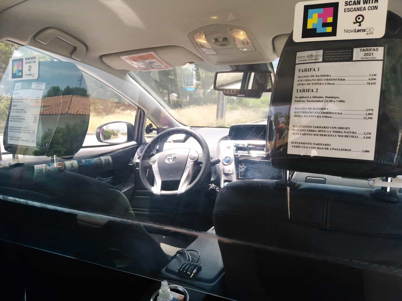 Veduta generale dell’interno di un taxi Toyota Prius di Benidorm dal sedile posteriore: volante, paratia, tariffario 2021 e cartello NaviLens GO in alto.