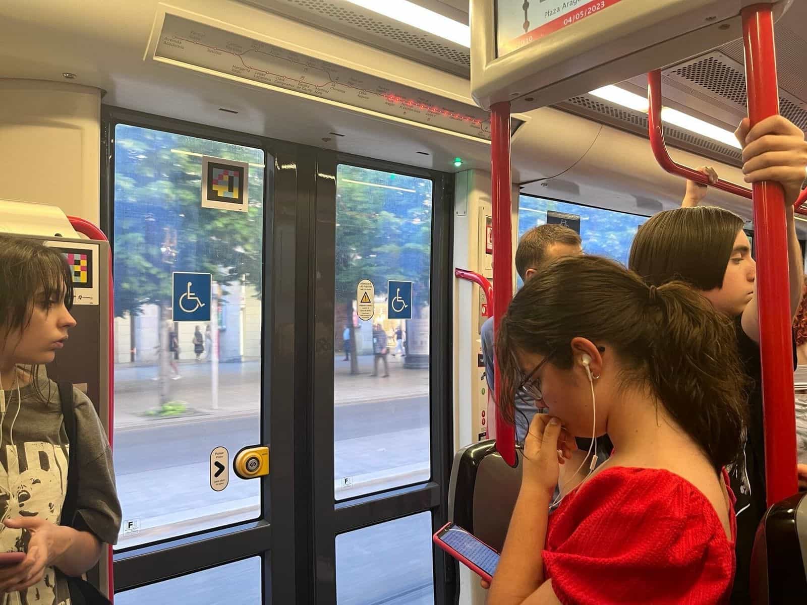 Interno di un tram di Saragozza in marcia: viaggiatori in piedi aggrappati alle barre rosse, pannello led superiore con la sequenza delle fermate illuminata fino a «Plaza Aragón 04/05/2023» e un codice NaviLens applicato sulla porta a vetri al centro della vettura, accanto ai pittogrammi del posto per sedia a rotelle