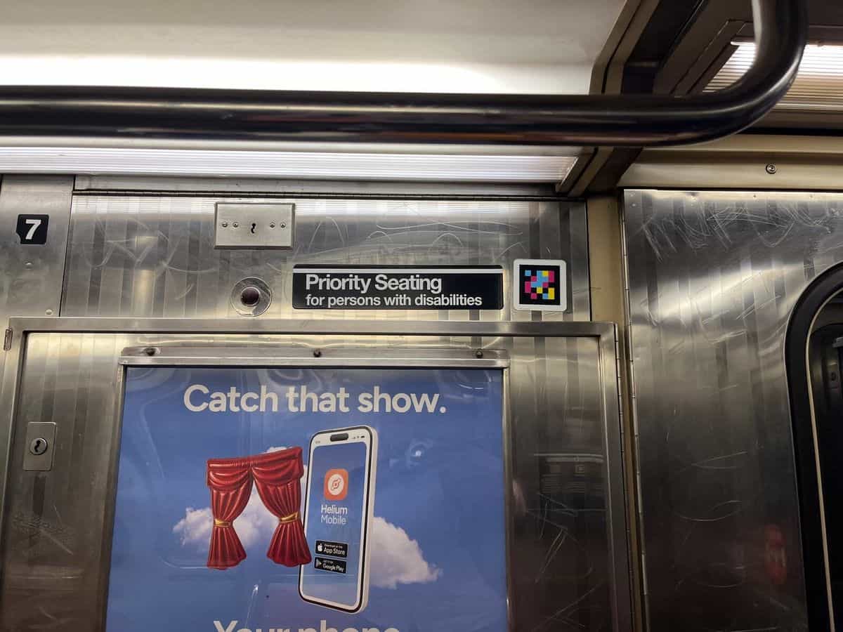 Detalle del interior de un vagón del subway de NY: cartel negro con texto blanco «Priority Seating for persons with disabilities», botón de emergencia rojo y, justo a la derecha, un código NaviLens de cuadros de colores enmarcado en blanco