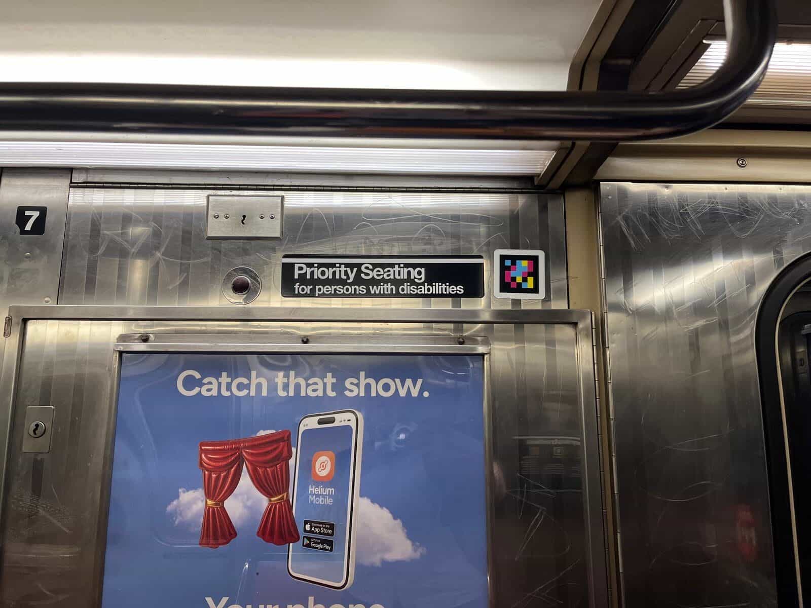 Detail im Inneren eines NYC-Wagens: schwarzes Schild mit weißem Text «Priority Seating for persons with disabilities», roter Notruf-Knopf und direkt daneben ein in Weiß gerahmter NaviLens-Farbcode