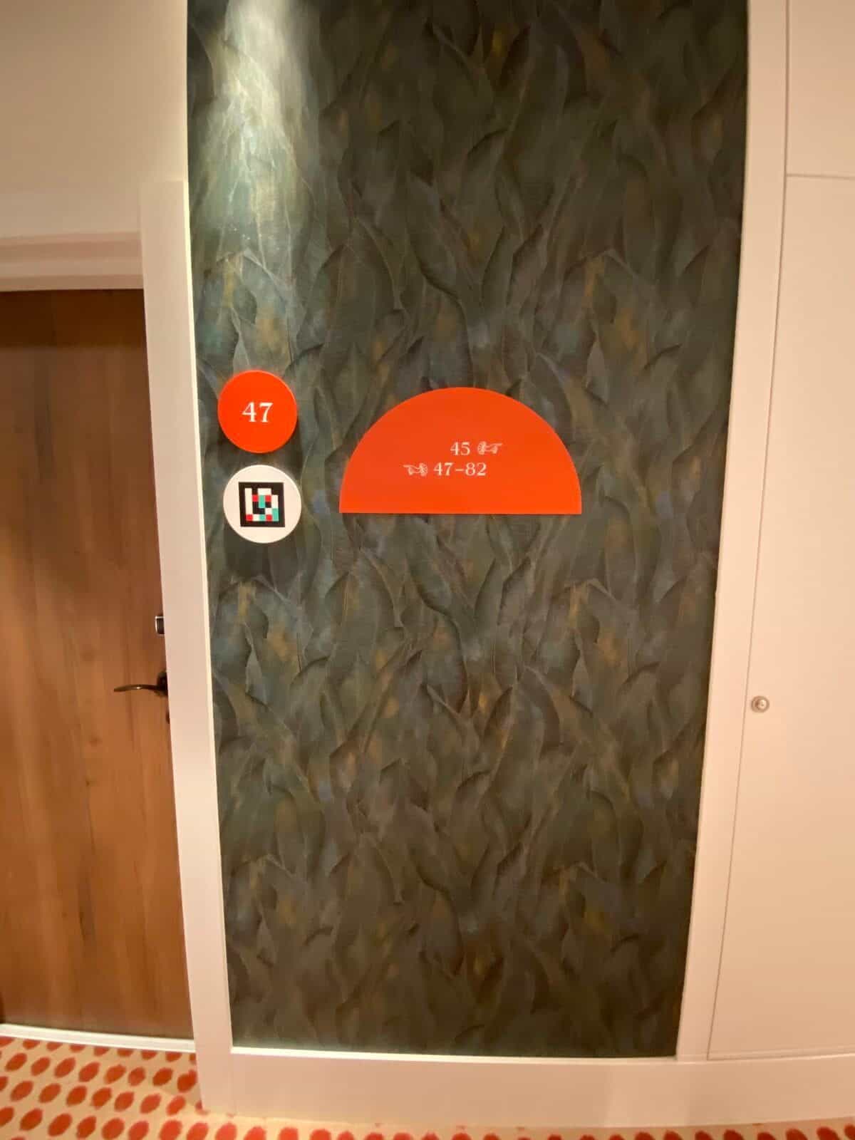 Türrahmen von Zimmer 47 am ibis Styles Sevilla auf grüner Tropentapete: oranger Disc mit „47“ und darunter oranger Halbmond „45 · 47-82“ mit Pfeilen und NaviLens-Code