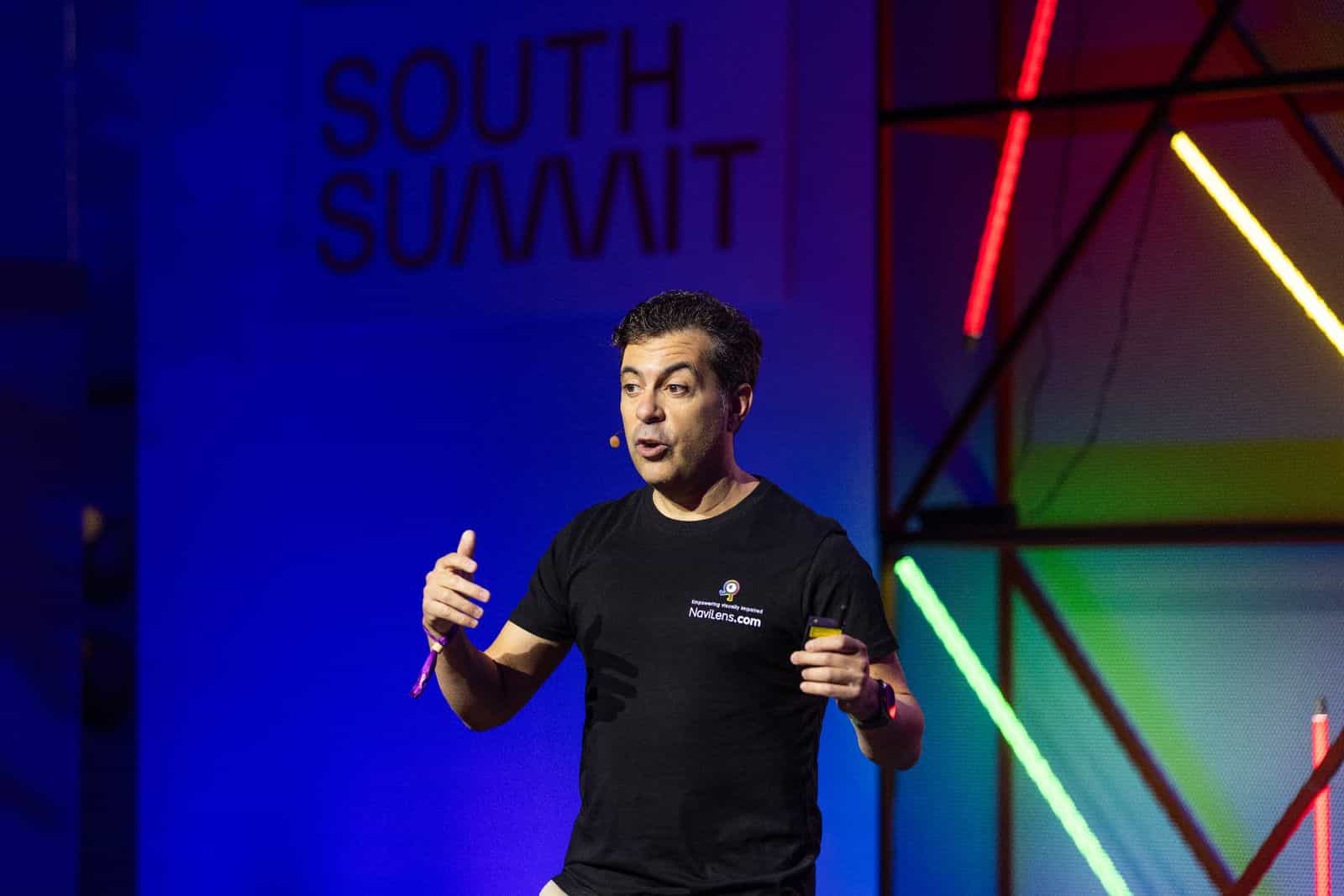 Javier Pita, CEO di NaviLens, sul palco del South Summit Madrid 2024 con una maglietta nera stampata con il logo di NaviLens e il motto «Empowering visually impaired — NaviLens.com»; in fondo, fondale blu intenso con la scritta «SOUTH SUMMIT» e barre di luce verde, giallo e rosso della scenografia del Mobility Stage