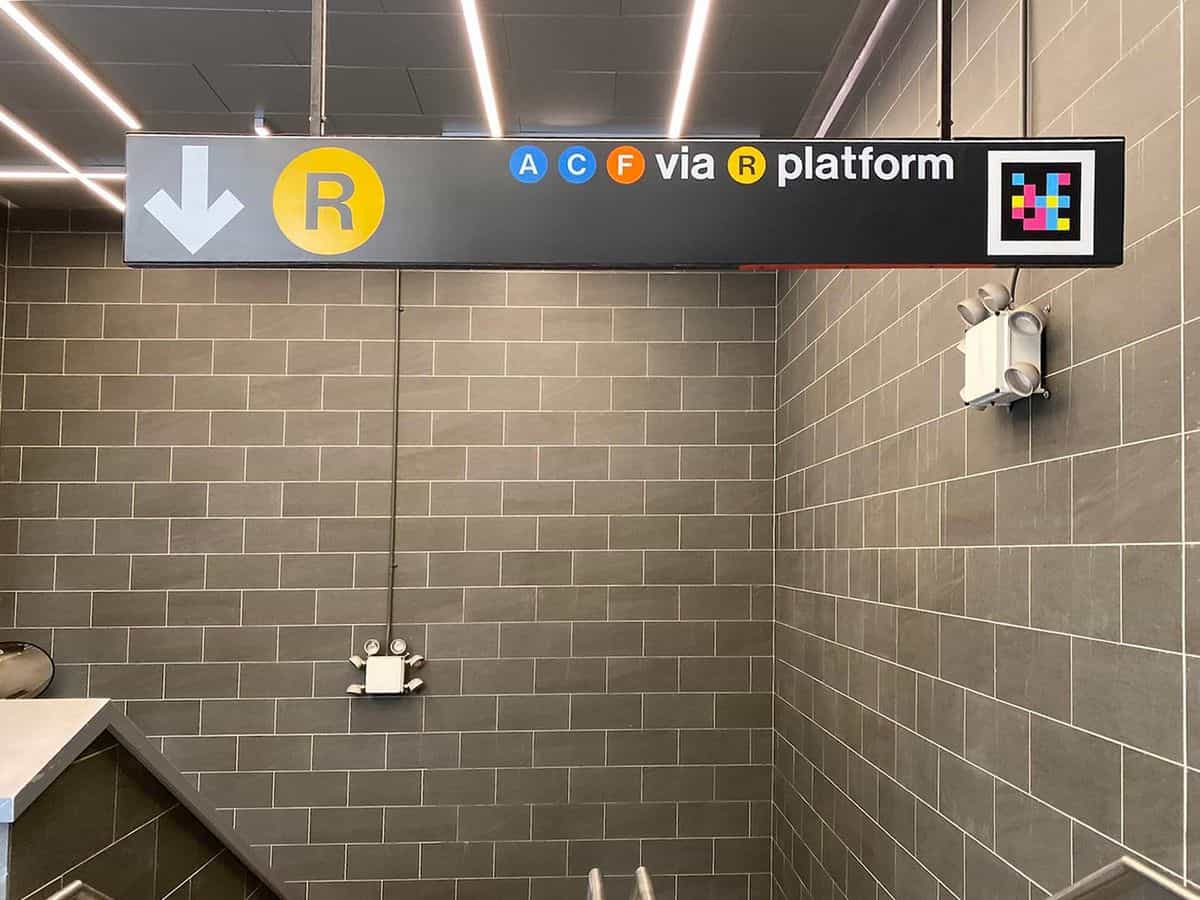 Cartel suspendido del techo en el vestíbulo de Jay St-MetroTech sobre paredes de azulejo gris: flecha blanca a la izquierda con el bullet amarillo R y, a la derecha, los bullets A C F con el texto «via R platform» y un código NaviLens cuadrado integrado en el extremo derecho del propio cartel oficial