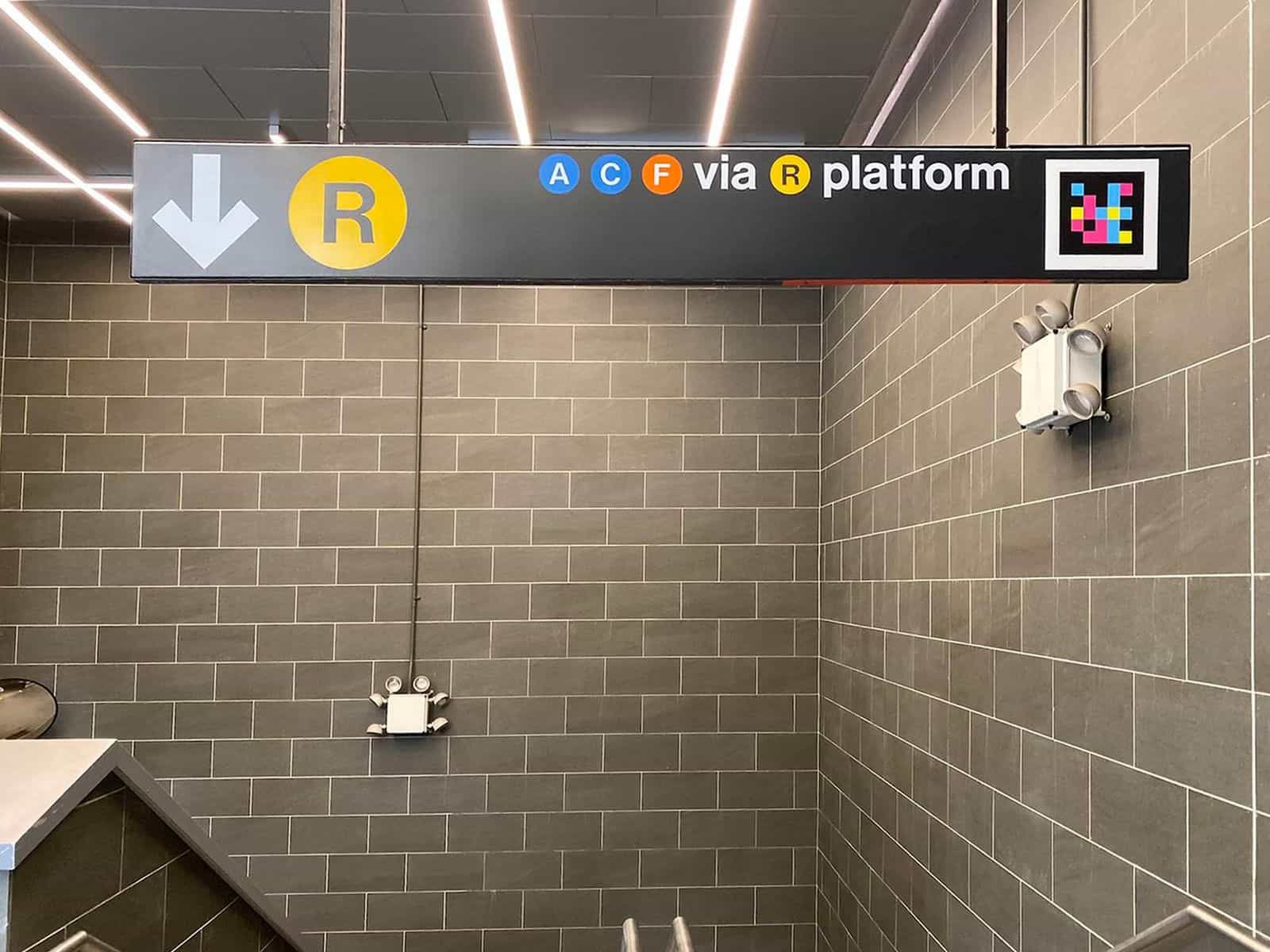 Ceiling-hung sign in the Jay St-MetroTech mezzanine over grey-tiled walls: white arrow to the left with the yellow R bullet and, to the right, the A C F bullets with the text «via R platform» and a NaviLens code embedded on the right edge of the official sign itself