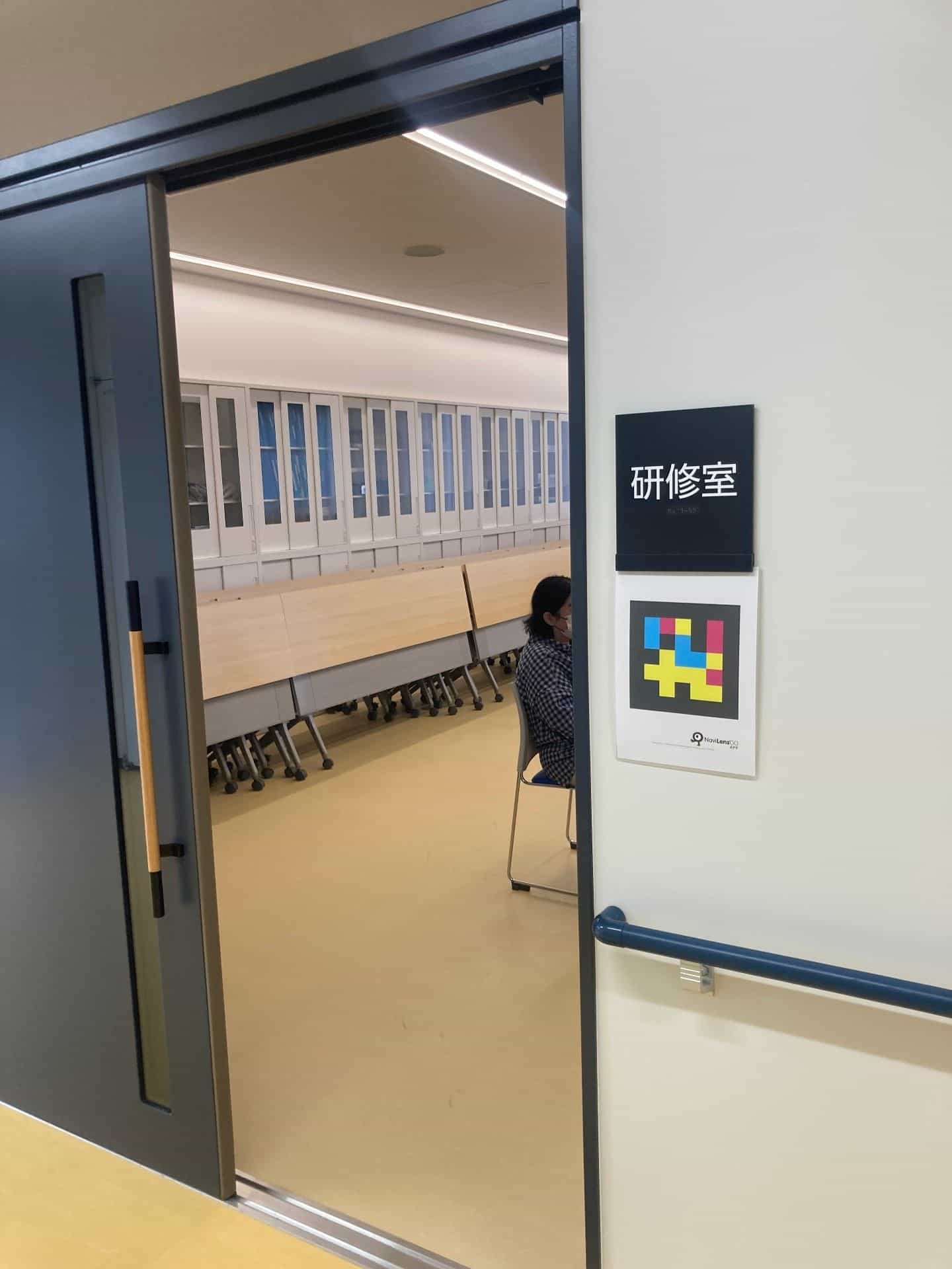 Porta scorrevole scura aperta dell’aula formazione («研修室») del centro: all’interno si vedono file di tavoli pieghevoli con sedie impilabili e, in fondo, una parete di armadi con vetrine di vetro bianco; sulla parete destra, targa tattile nera con il testo bianco «研修室» e, sotto, un cartello A4 con il codice NaviLens GO in ciano, magenta e giallo su fondo nero; corrimano tubolare blu e pavimento in vinile giallo